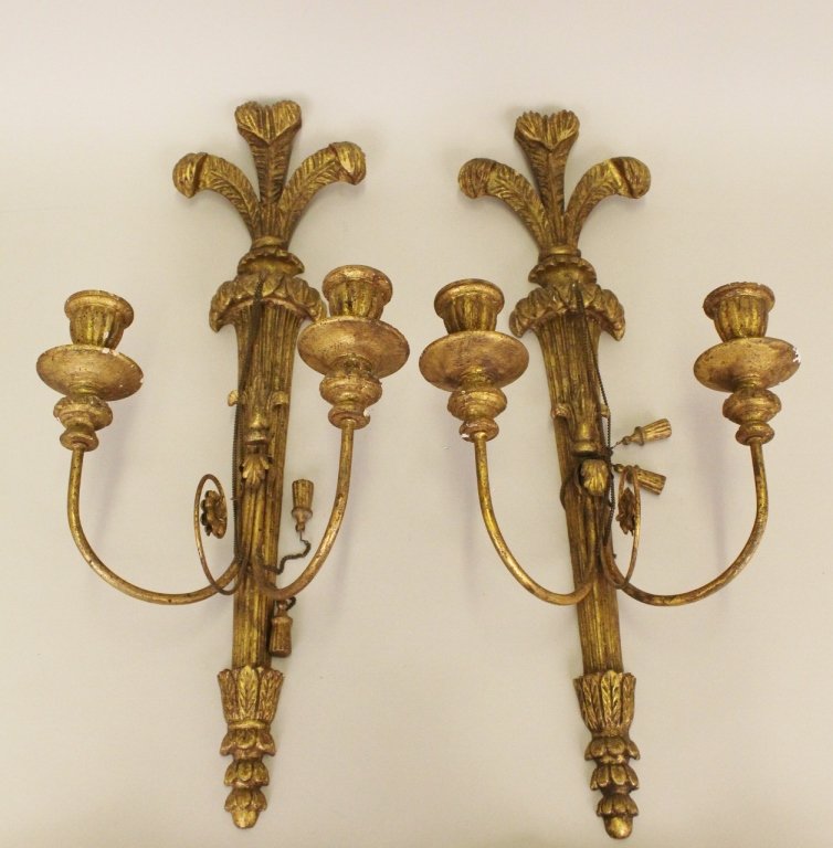 A Pair of Italian Gilt Wood Wall Sconces: With two-arms terminating in candleholders and foliate decoration. Two tassels hang at middle from dangling chains. With Italian label on verso. 26 inches x 11 inches.
