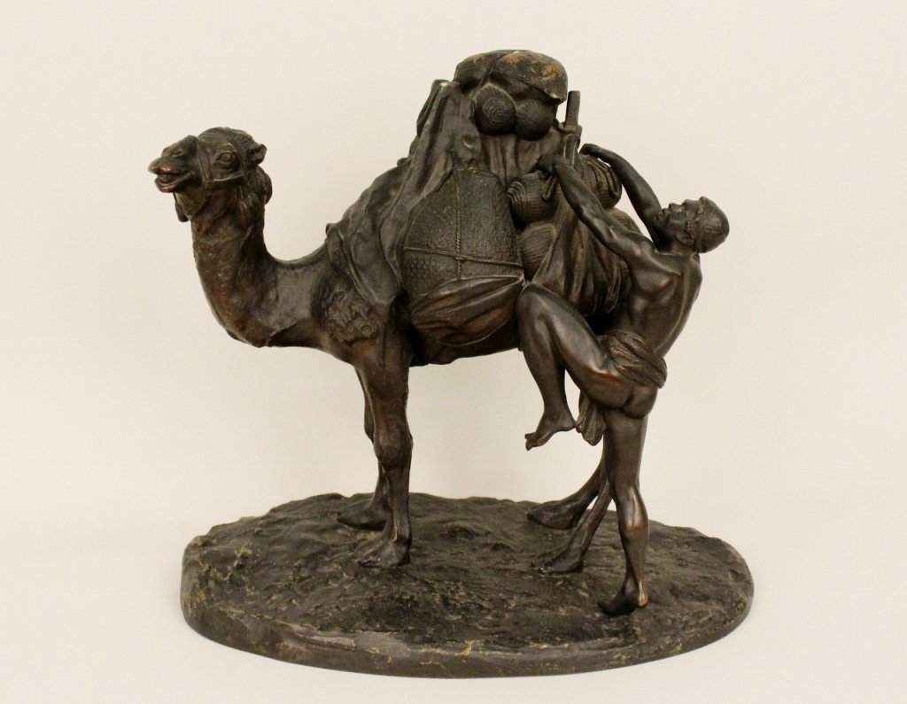 Orientalist Bronze of Figure with Camel: Finely rendered Orientalist-themed patinated bronze figure of a large camel with a tasseled headdress carrying large roped baskets and being tended to by a young semi-nude man standing on tip toes wit