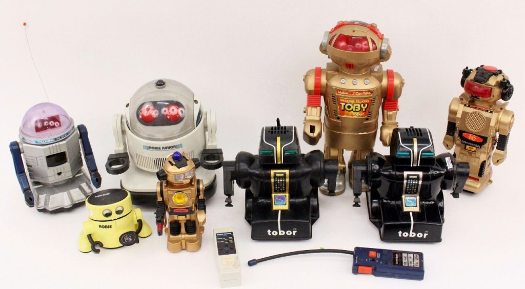 Group of Eight Robots, Circa 1980s: Group of eight robots (circa 1980s) including two black tobor robots; a walking talking toby robot in gold and red tones; a gold tone Pyrobot; a 2002 Model 2 gold tone robot; a Radio Shack Robie Junio