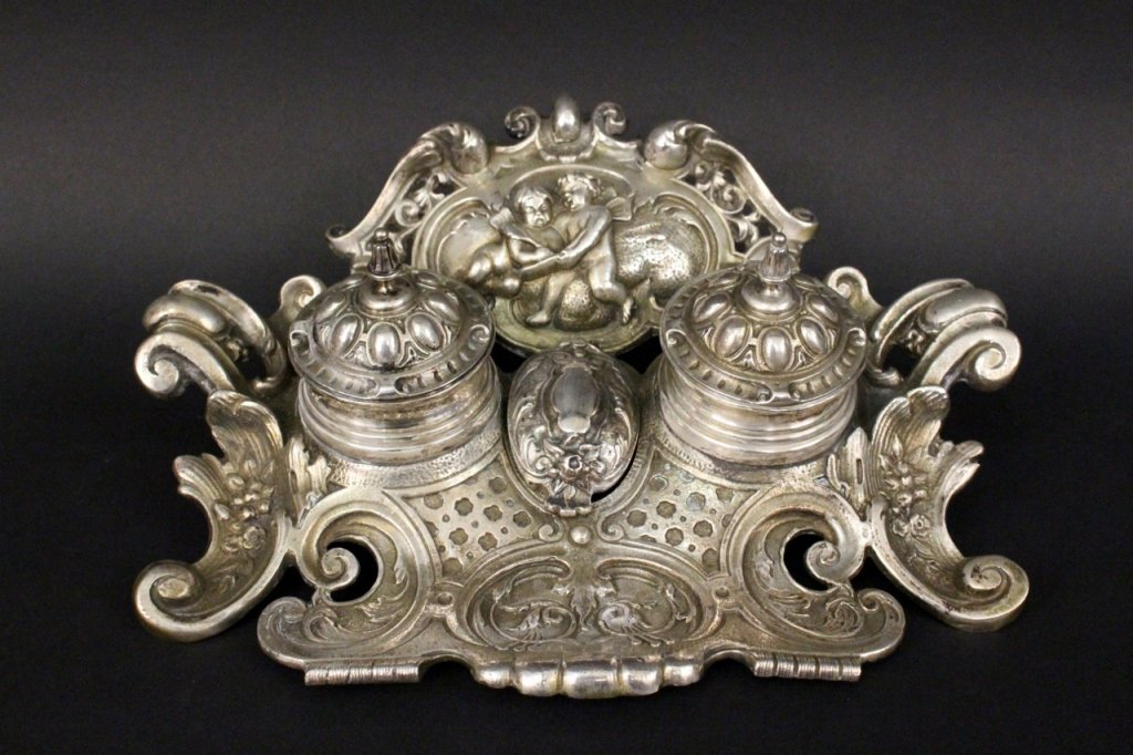 800 Silver Figural Inkwell, Marked: Continental School, 19th century. 800 silver figural inkwell with two winged cherubs writing in a book with a feathered quill in medallion at top above lidded inkwells with glass inserts toped with po