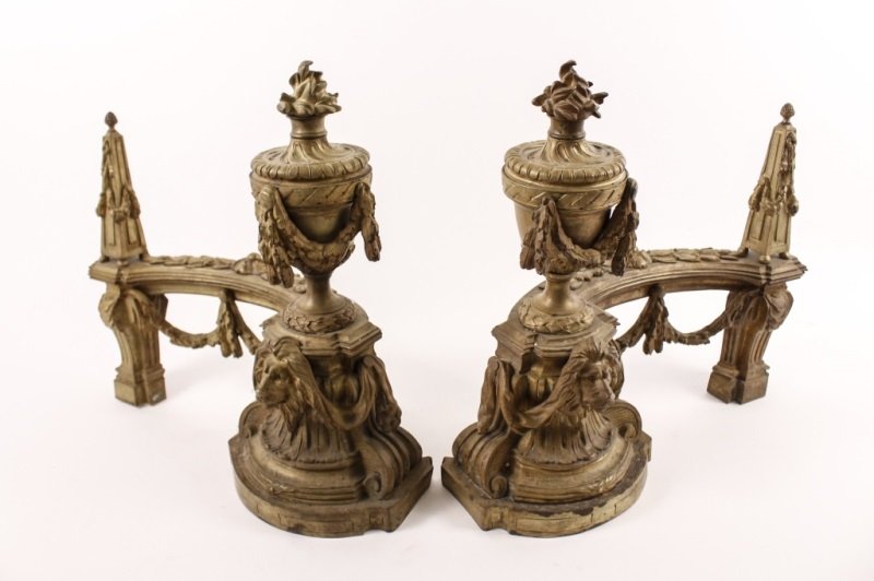 Pair of Louis XVI Style Gilt Bronze Chenets: French, Late 19th Century. Pair of Louis XVI style gilt bronze chenets, decorated with lion's heads on the bases, and swagged urn finials trimmed in egg and dart borders, capped with flambeau finials.