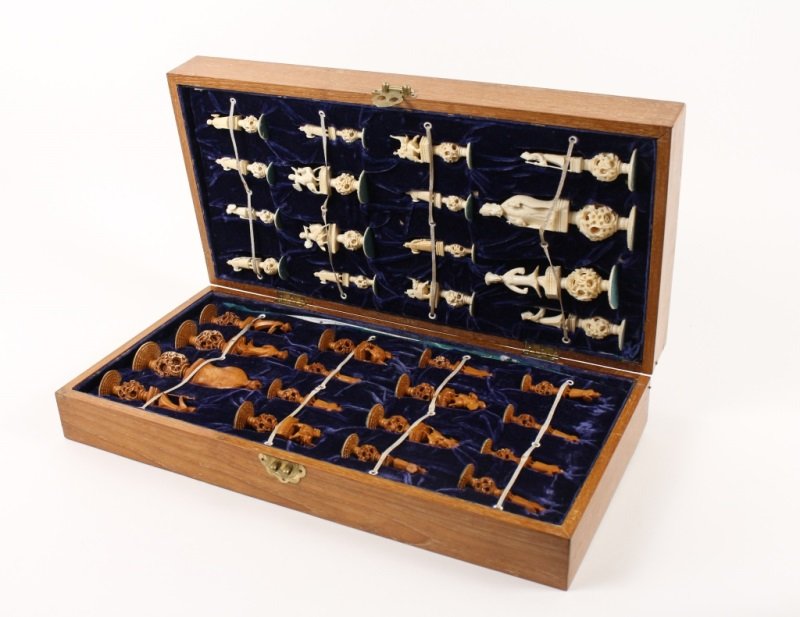 Chinese Carved Ivory Chess Set w/ Board: Chinese, Early 20th Century. Chinese carved ivory chess set including a wooden case with chess board top, holding 32 carved ivory figural pieces, 16 of which are tinted, all raised on puzzle ball base