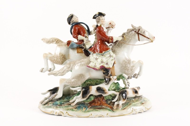 Early 20th C. German Porcelain Fox Hunt Scene: German, late 19th/ early 20th century. A figural porcelain sculpture depicting a fox hunt with a male and female rider each on horseback and wearing red long coats above floral dress and/or blouse, wi