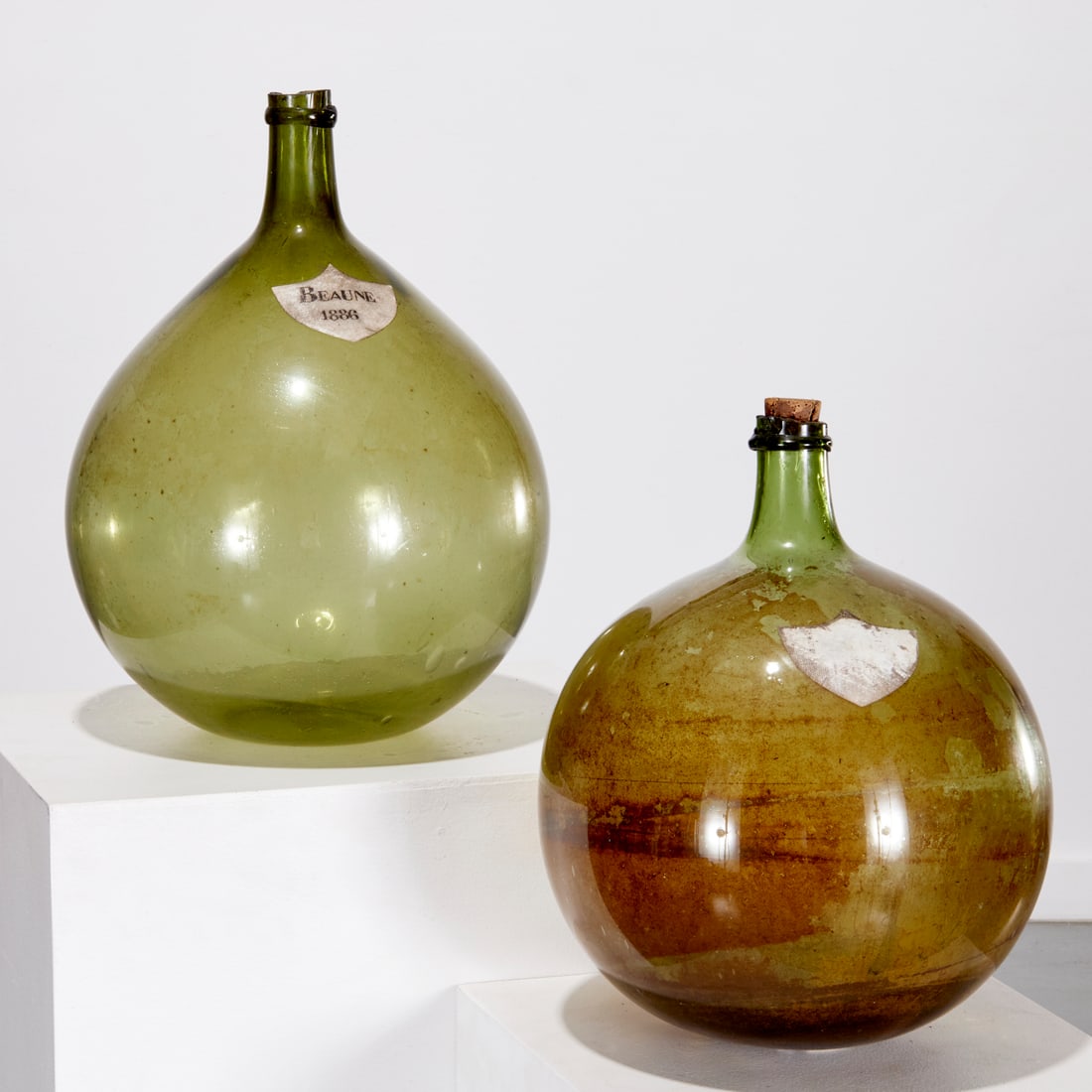 (2) Antique French blown glass wine carboys: (2) Antique French blown glass wine carboys, 19th c., one with shield form label "Beaune, 1886", the other with remnant label with wording worn off, 20"h x 15"w (larger), 17"h x 13"w (smaller)