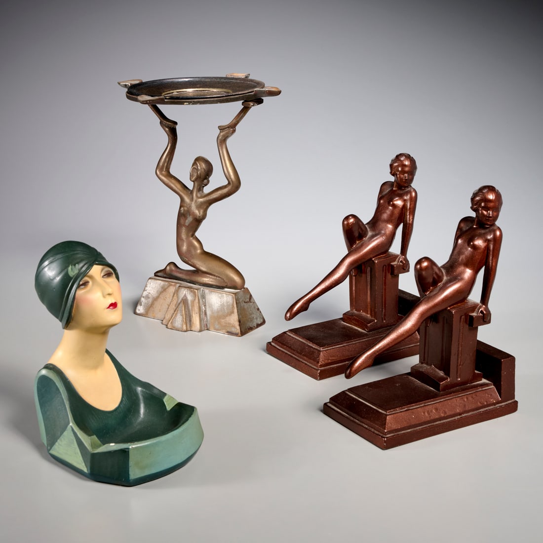 Group (4) Art Deco figural decorations: Group (4) Art Deco figural decorations, 20th c., incl. a pair of patinated metal female nude bookends, a painted ceramic woman ashtray, and a patinated metal kneeling female nude ashtray, with