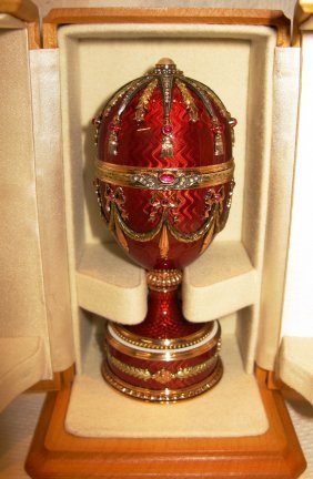 RUSSIAN JEWELED GOLD GUILLOCHE EGG: the ovoid body having a ground of semi-translucent ruby moire enamel with applied ribbon-tied, ruby-set, two-color gold foliate swags. The cover set with adularescent moonstone, seed pearls, rose cut