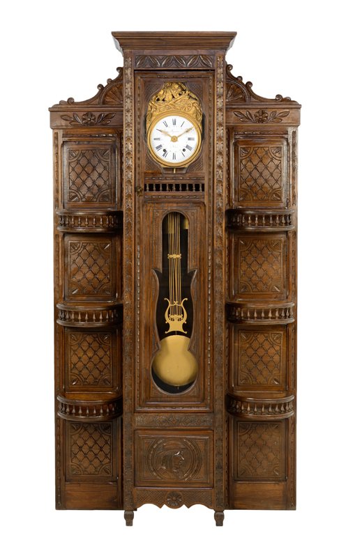 A FRENCH NORMAN BRITTANY LONGCASE PENDULUM CLOCK WITH: A FRENCH NORMAN BRITTANY LONGCASE PENDULUM CLOCK WITH SHELVING Early-Mid 20th Century A carved Brittany pendulum clock features a harvest themed repousse style clock face with enamel painted numerals,