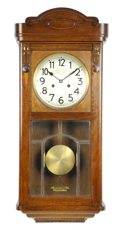 AN ART DECO STYLE WALNUT REGULATOR WALL CLOCK: AN ART DECO STYLE WALNUT REGULATOR WALL CLOCK The clock with a domed cornice having a round clockface flanked by a carved cartouche above a glaze panel revealing the brass pendulum, ending in a taperi