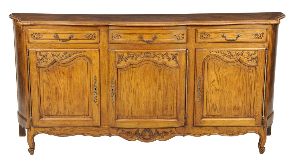 A FRENCH PROVINCIAL STYLE OAK SIDEBOARD: A FRENCH PROVINCIAL STYLE OAK SIDEBOARD The sideboard having a shaped rectangular top over three short drawers and three paneled doors, above the serpentine apron centered with a carved shell, raised 