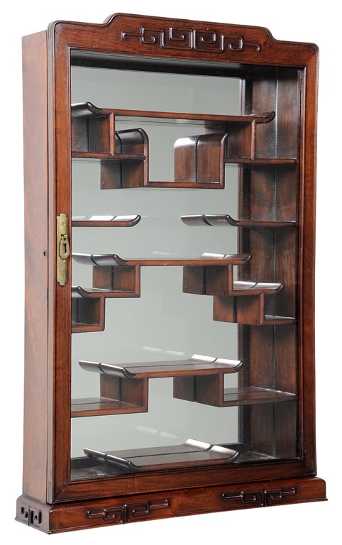 A CHINESE HARDWOOD WALL VITRINE: A CHINESE HARDWOOD WALL VITRINE The carved hardwood wall vitrine having a shaped pediment with Greek key motif, above the conforming case, the single door having brass pull and mounts and opening to t