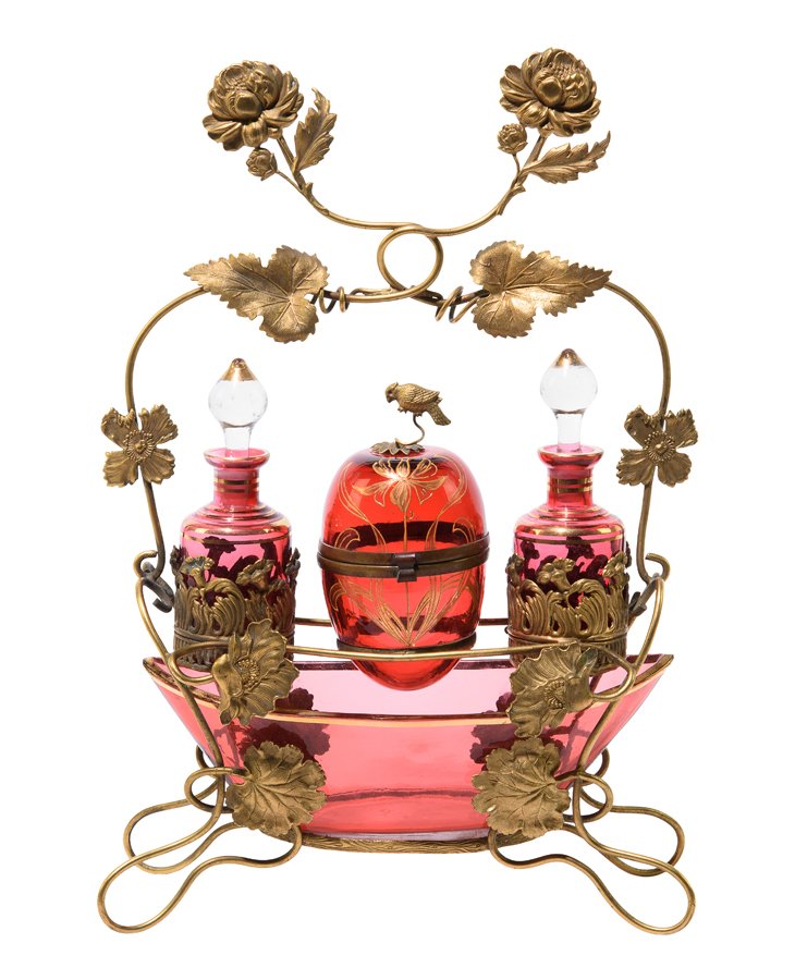 AN ART NOUVEAU CRANBERRY GLASS DRESSER SET, IN THE: AN ART NOUVEAU CRANBERRY GLASS DRESSER SET, IN THE MANNER OF MOSER Early 20th century The gilt metal stand having foliate and scrolling ivy leaf decoration, fitted with an oval cranberry glass bowl, t