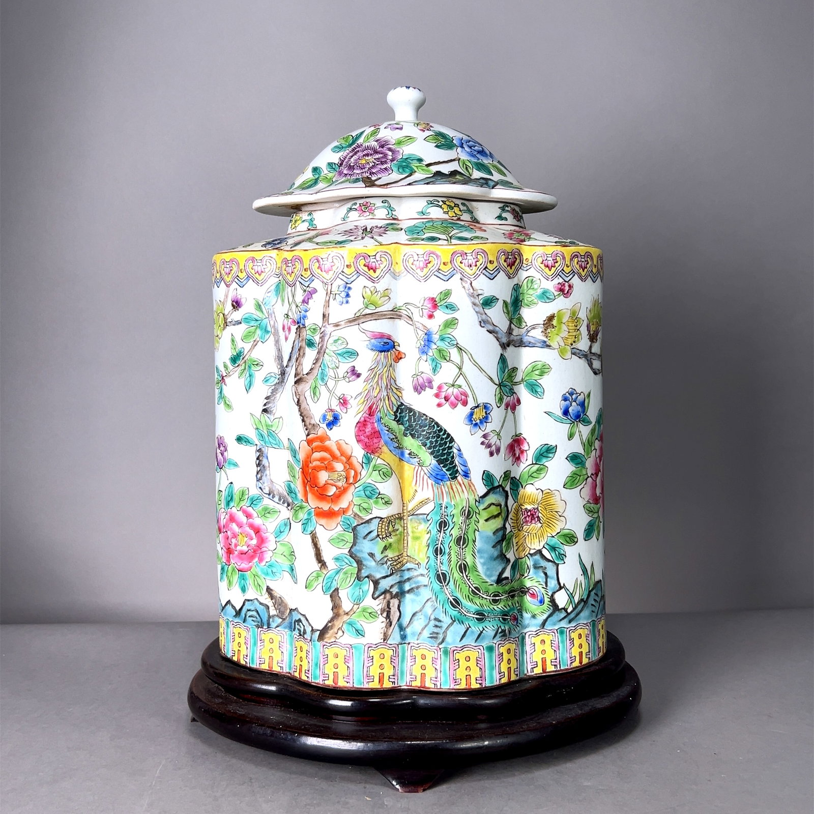 Chinese Famille Rose Lidded Porcelain Jar: Chinese famille rose porcelain lobed jar with a lid, featuring hand-painted scenes of peacocks, vibrant florals, and intricate patterns, on wooden stand. Dimensions: Height: 14 in. Width