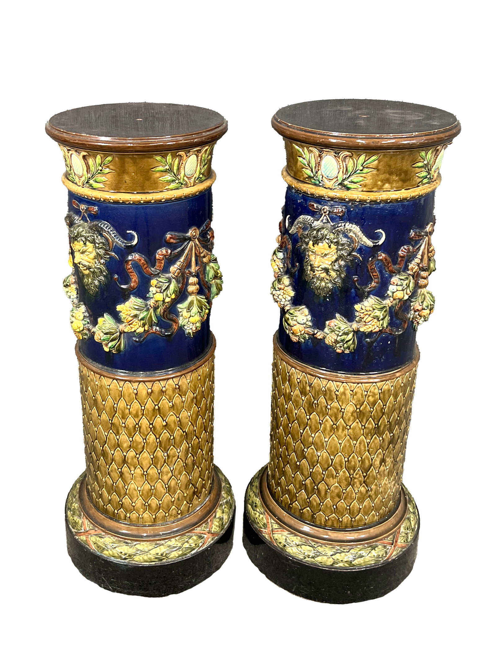Pair of Majolica Pedestals: Pair of majolica pedestals with intricate detailing, featuring raised floral and foliage motifs, along with a decorative satyr mask. The body of the pedestal is adorned with basket-weave and latt