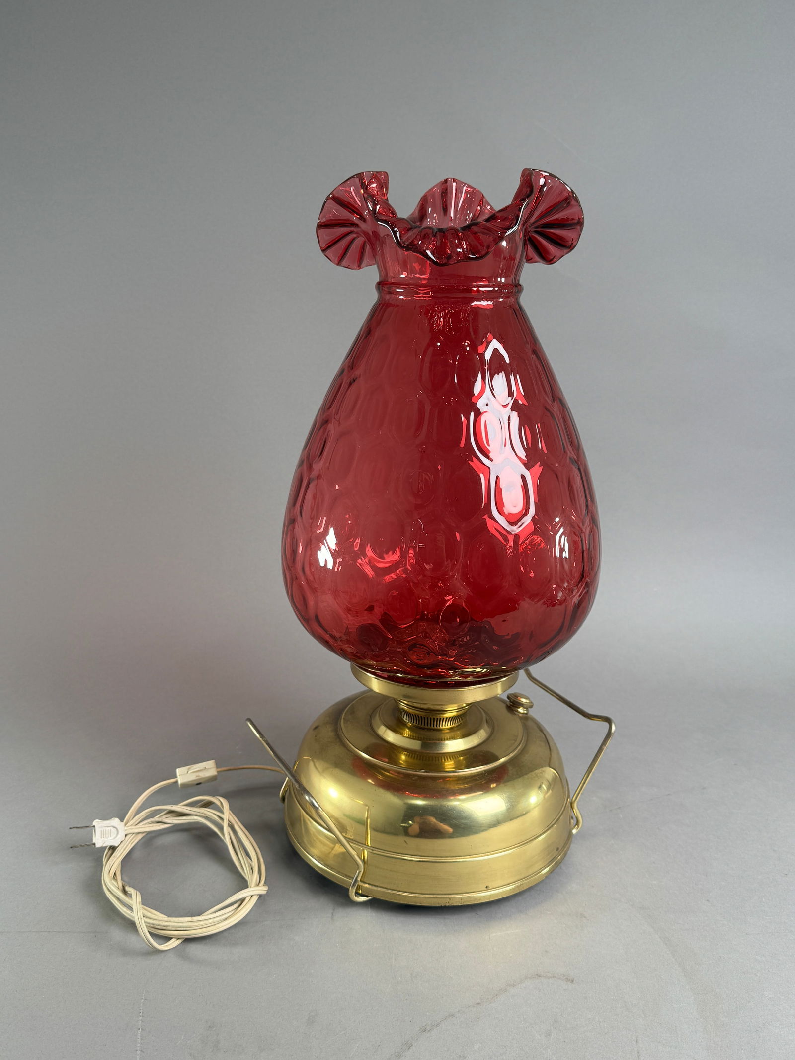 Vintage Cranberry Glass Table Lamp: Vintage table lamp featuring a cranberry glass shade with a ruffled edge and textured pattern. The shade is mounted on a brass-toned fixture with electrical wiring. Measures approximately 19 inches ta