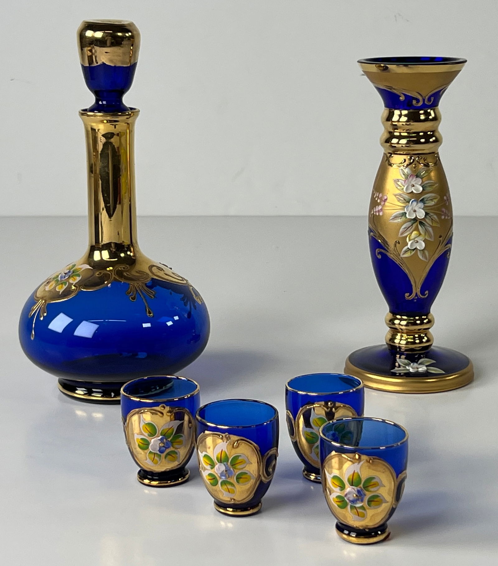 Italian Cobalt Blue Glass Decanter Set with Candlestick: Cobalt blue glass decanter set with gold gilt floral decoration. Includes a decanter with stopper, four shot glasses, and a candlestick holder. Marked 'Made in Italy'. Height 9 1/2 inches.