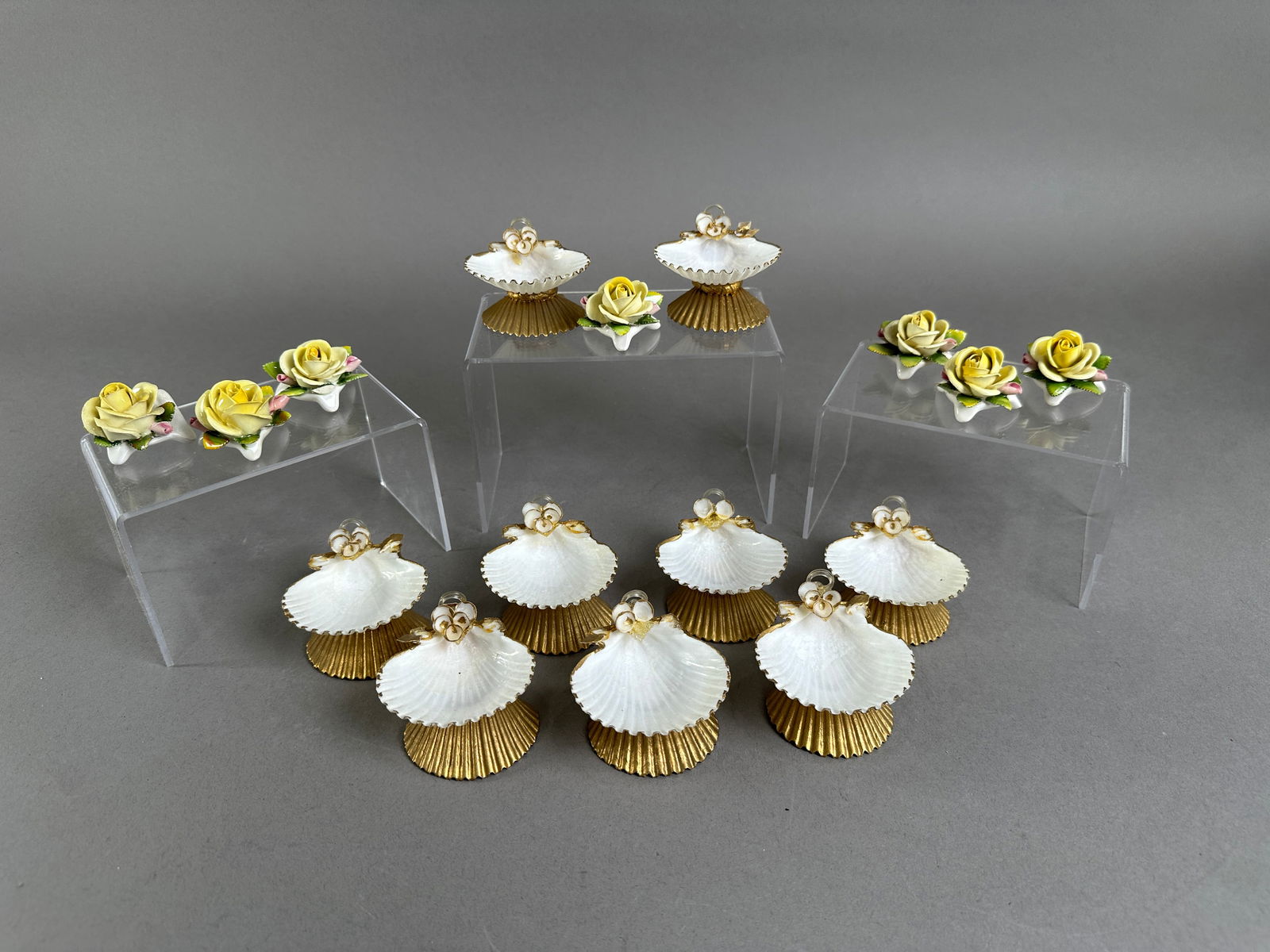 Collection of Porcelain Shell and Rose Place Card Holders: Collection of porcelain place card holders, including 9 shell-shaped holders with gold bases and floral accents, and 7 rose-shaped holders with yellow petals and green leaves. Size ranges from 1.5'' t