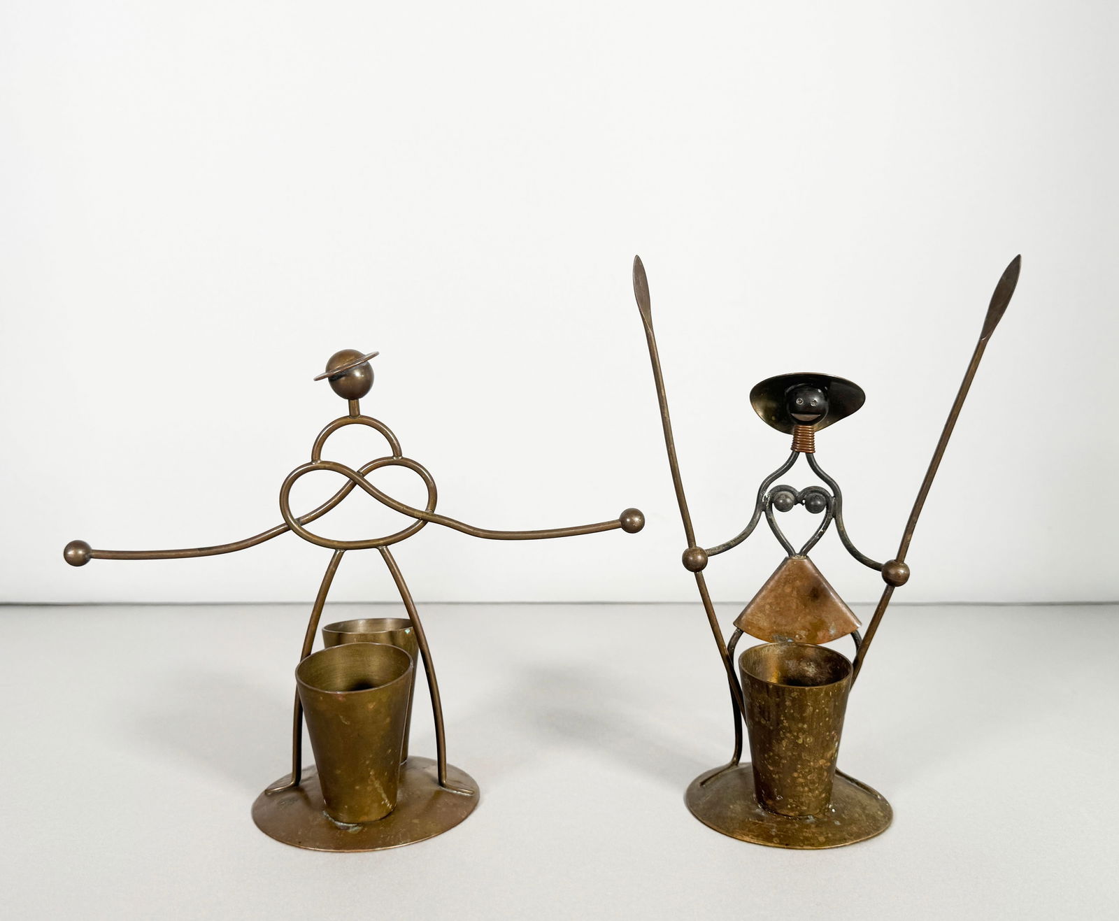 Mid-Century Modern Metal Sculptures: Pair of mid-century modern metal sculptures depicting stylized figures. Each figure holds a small brass cup. The sculptures are crafted from wire and metal. Dimensions: 7'' x 8.25'' 8'' x 5'' Weigh