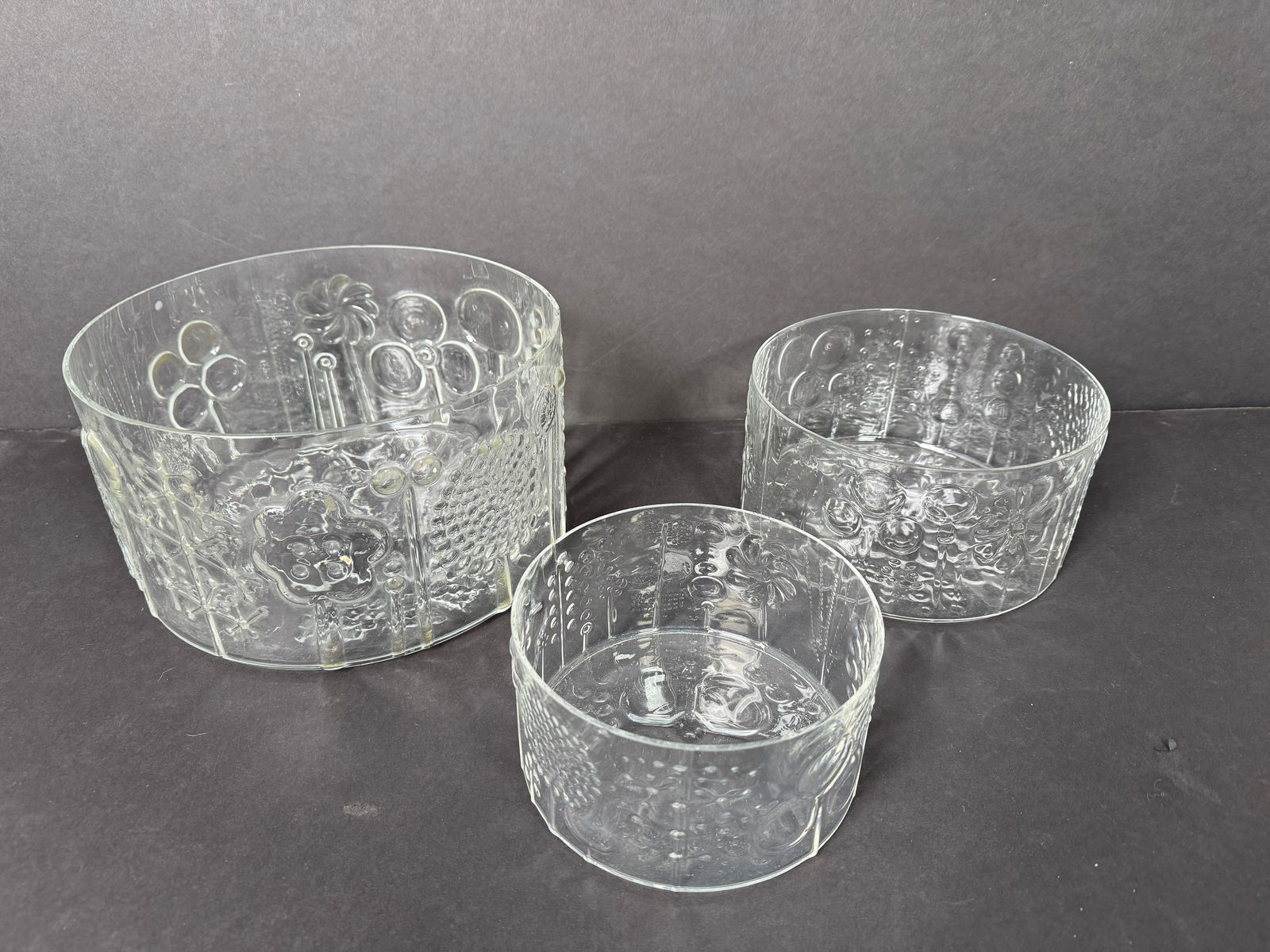 Three Iittala Flora Glass Bowls by Oiva Toikka: Set of three clear Iittala Flora glass bowls designed by Oiva Toikka. Each bowl features a unique embossed floral design. Diameters: 6", 7.5", 9.5"