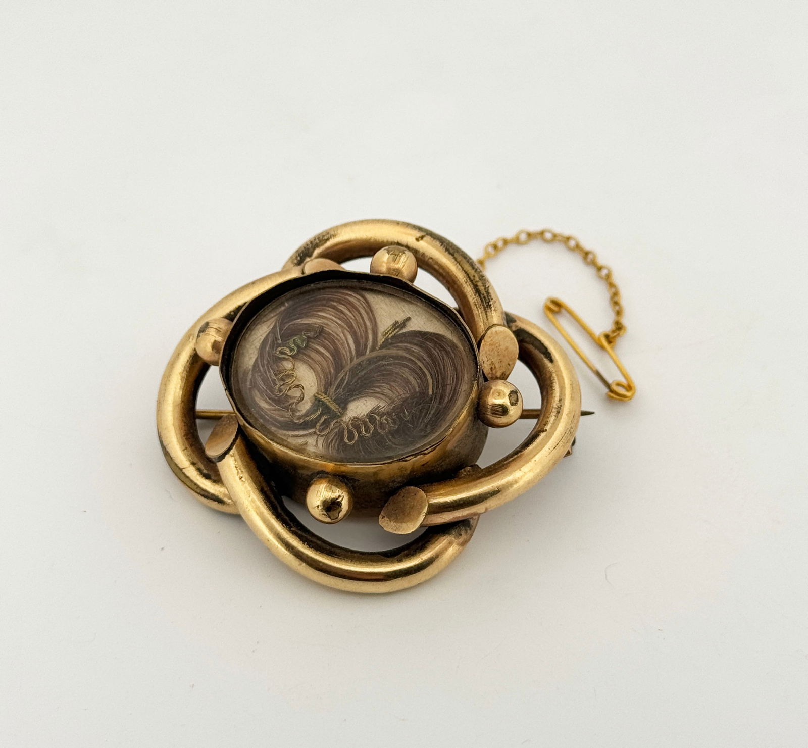 Victorian Mourning Hair Art Brooch: Victorian era mourning brooch featuring hair art encased in a gold-tone metal frame. The brooch has a safety chain and pin. Measures approximately 1 3/4 x 1 5/8 inches.21.2g