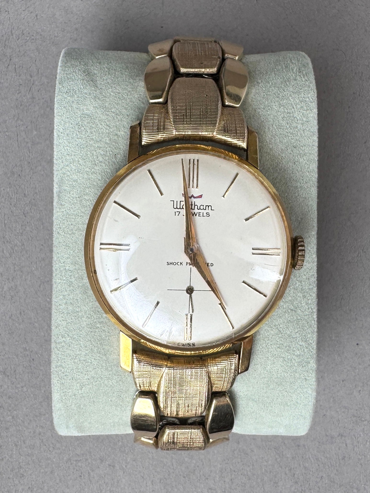 Waltham 17 Jewels Gold Tone Wristwatch: Waltham gold tone wristwatch. Features a white face with gold tone hands and markers. Marked 'Waltham 17 Jewels Shock Protected'. Stainless steel back. Swiss made. Unbreakable mainspring.