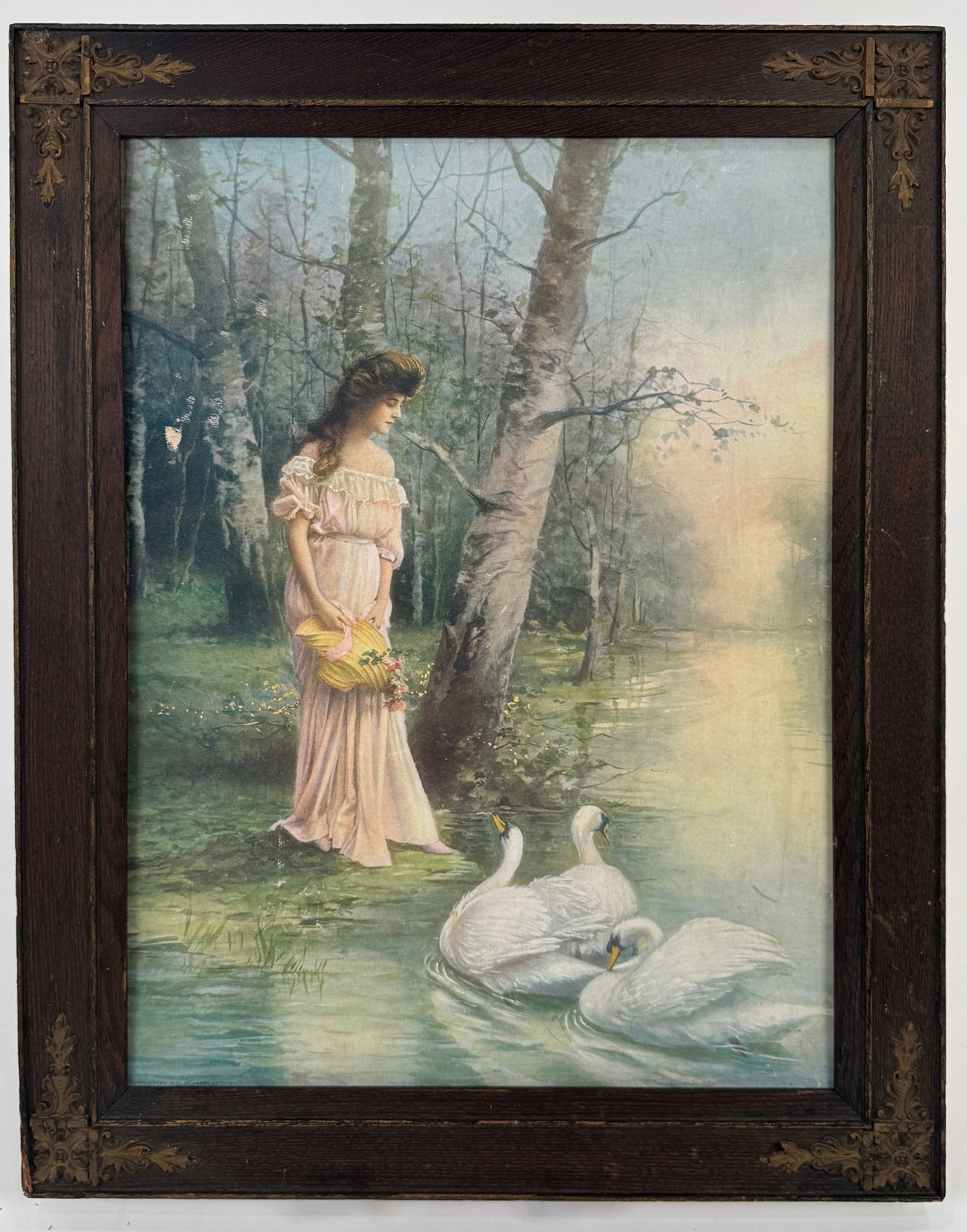 Framed Print: Woman with Swans by Lake - James Arthur: Framed print depicting a woman in a pink dress standing near a lake with swans. Copyrighted 1903 by James Arthur. The print is set in a decorative wooden frame. Frame measures 19.25 x 15.25 inches;