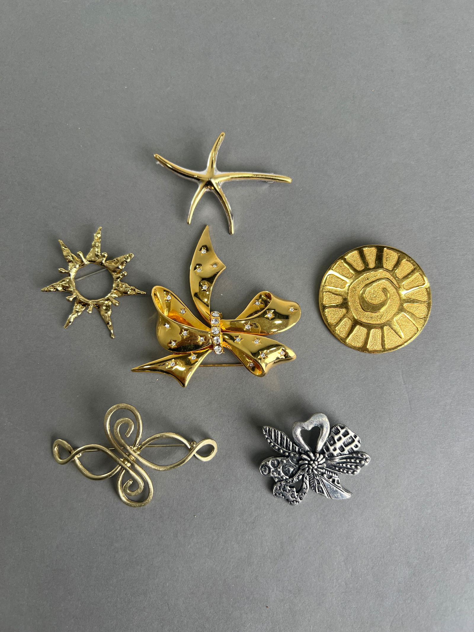Collection of Six Costume Brooches: A collection of six costume brooches, featuring a variety of designs including a sun, starfish, bow, and floral motif. The brooches are crafted from gold-tone and silver-tone metal. Dimensions vary.