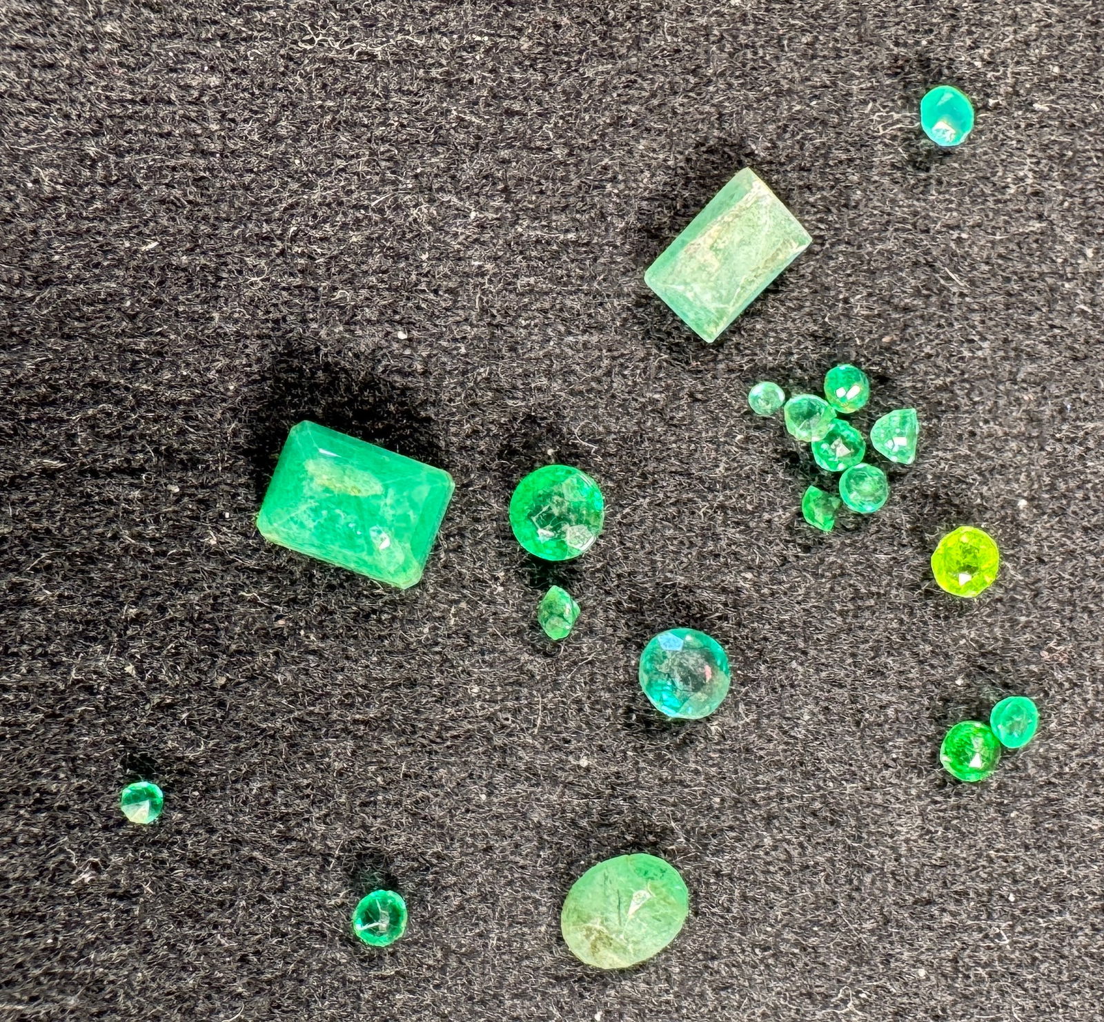 Loose Emerald Gemstones: Collection of loose emerald gemstones. Total weight approximately 0.3 oz. Sizes vary, with the smaller measuring approximately 1/8 inch. Includes various cuts and shapes.