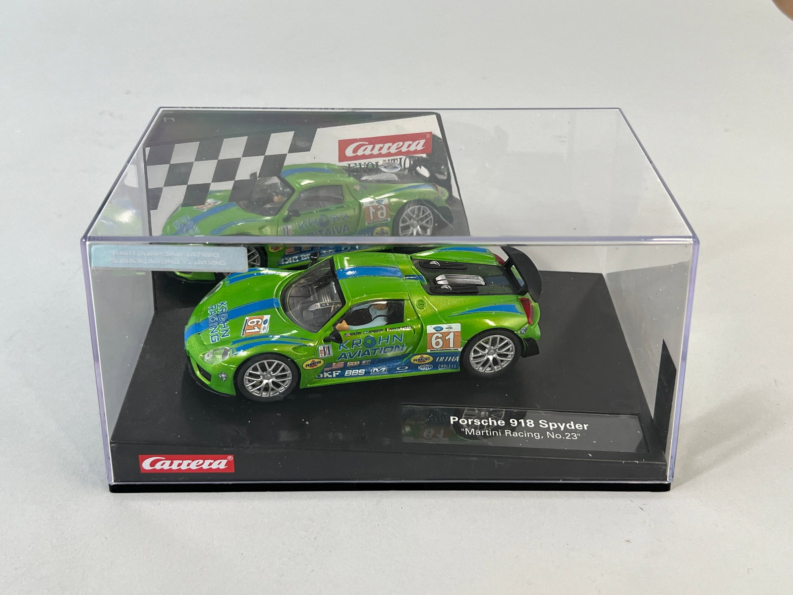 Carrera Porsche 918 Spyder #61: A green Carrera 1/32 slot car model of the Porsche 918 Spyder, featuring blue racing stripes and sponsor decals. The car is presented in its original display case with a checkered flag background.