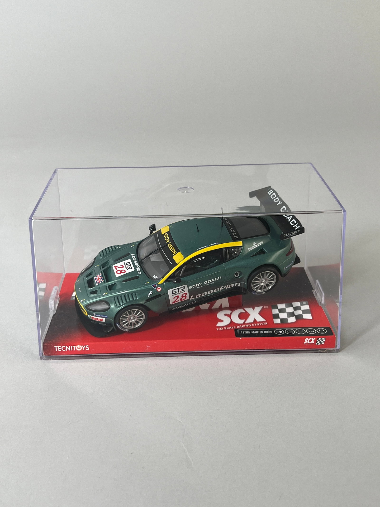 SCX Aston Martin DBR-9 #28: SCX Aston Martin DBR-9 #28 1/32 Slot Car. It is presented in its original clear display case with a red and black checkered base. Weight: 8oz. Dimensions: 4'' x 7'' x 3.5''