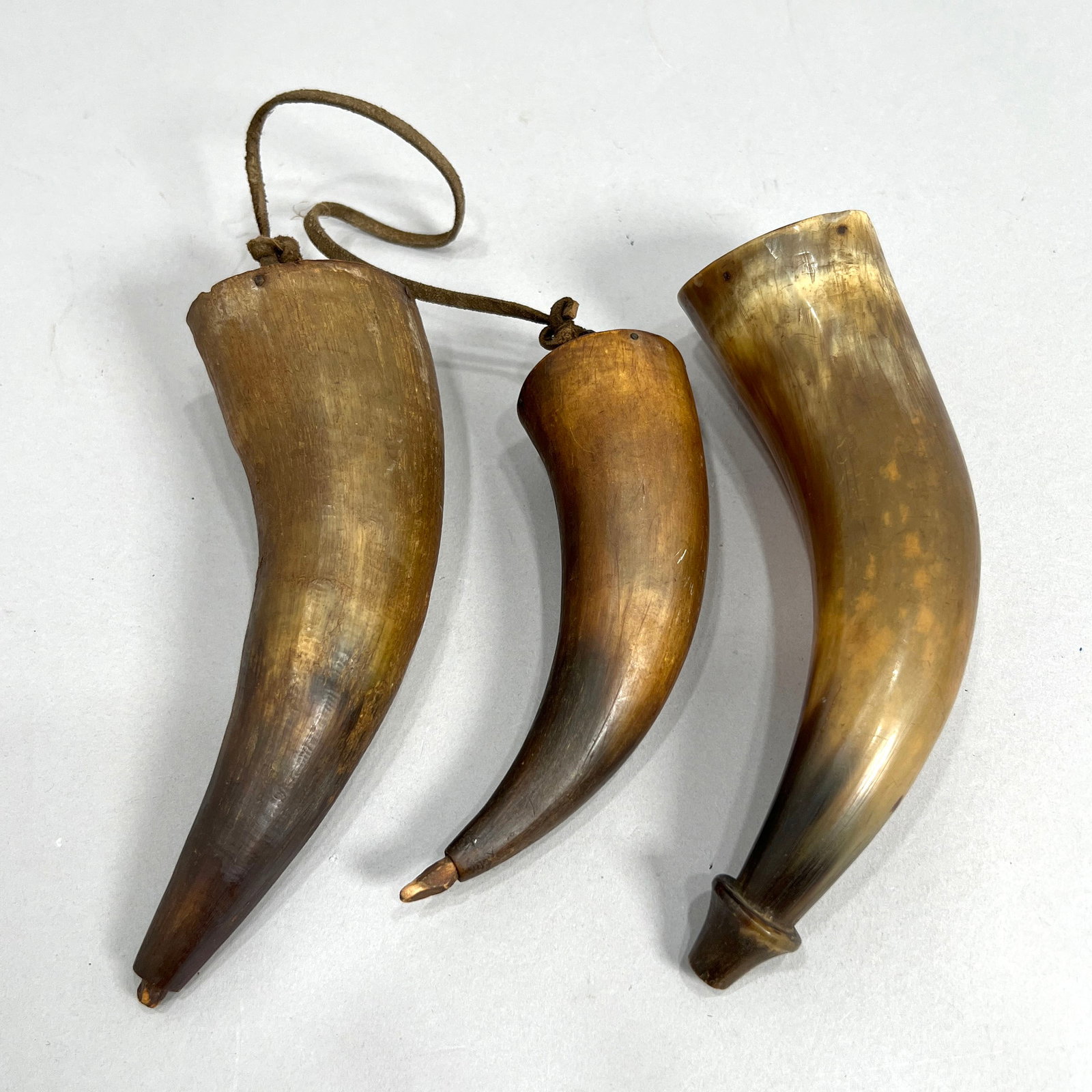Three Antique Powder Horns: A set of three antique powder horns with wooden stoppers. Maker's signature and date present on one stopper - see photo for details. The largest horn measures approximately 7.75 inches. The