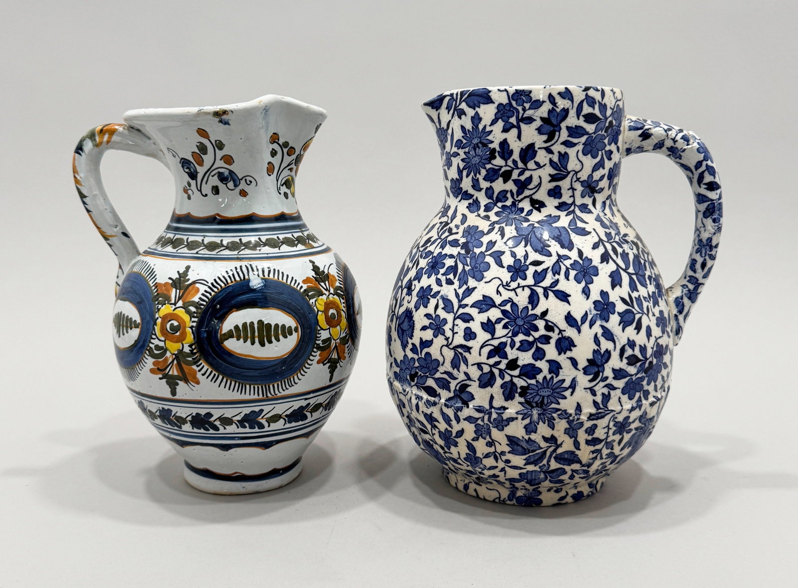 Two Floral Ceramic Pitchers: Two ceramic pitchers with blue, orange, and yellow floral designs. One is a hand painted Mexican Talavera pitcher, and the other features a blue transferware pattern. The hand painted pitcher is