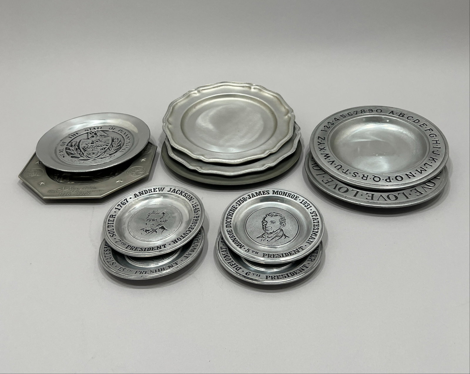 Collection of Pewter Plates and Platters: A collection of decorative pewter plates and platters, including a bicentennial commemorative plate depicting "A New Nation" and "We the People" text, and several plates featuring
