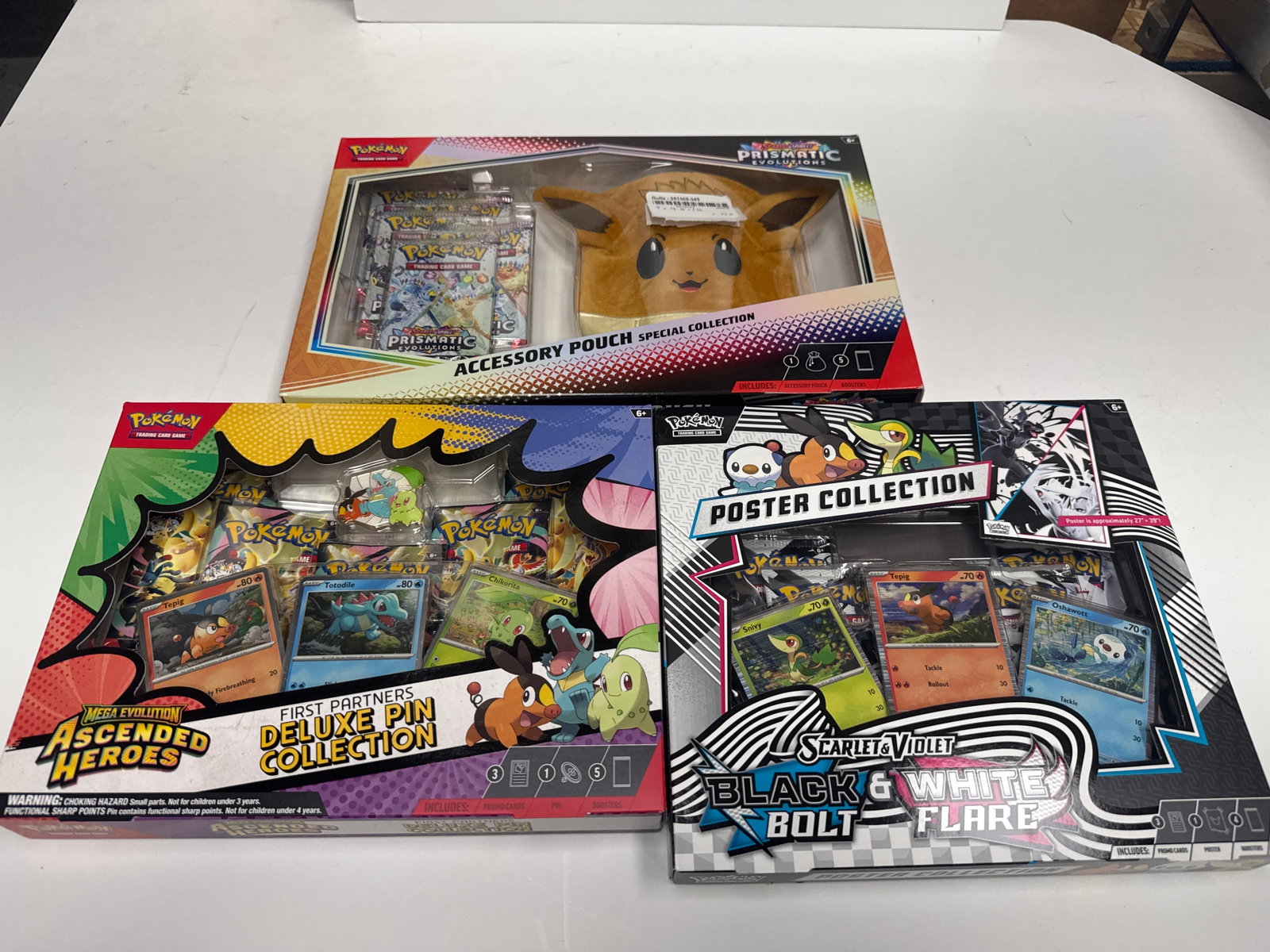 Pokemon TCG Accessory Pouch Special Collection, First Partners Deluxe Pin Collection, and Poster: This lot includes three Pokemon Trading Card Game (TCG) collections: the Prismatic Evolutions Accessory Pouch Special Collection featuring Eevee, the Mega Evolution Ascended Heroes First Partners Delu