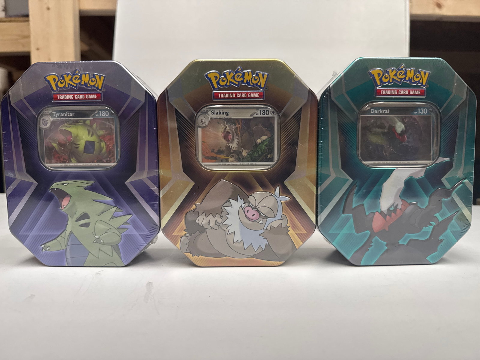 Pokemon Trading Card Game Tins - Tyranitar, Slaking, and Darkrai: This lot includes three Pokemon Trading Card Game tins featuring Tyranitar, Slaking, and Darkrai. Each tin contains 3 Pokemon TCG booster packs and a code card for Pokemon TCG Live. The Tyranitar tin 