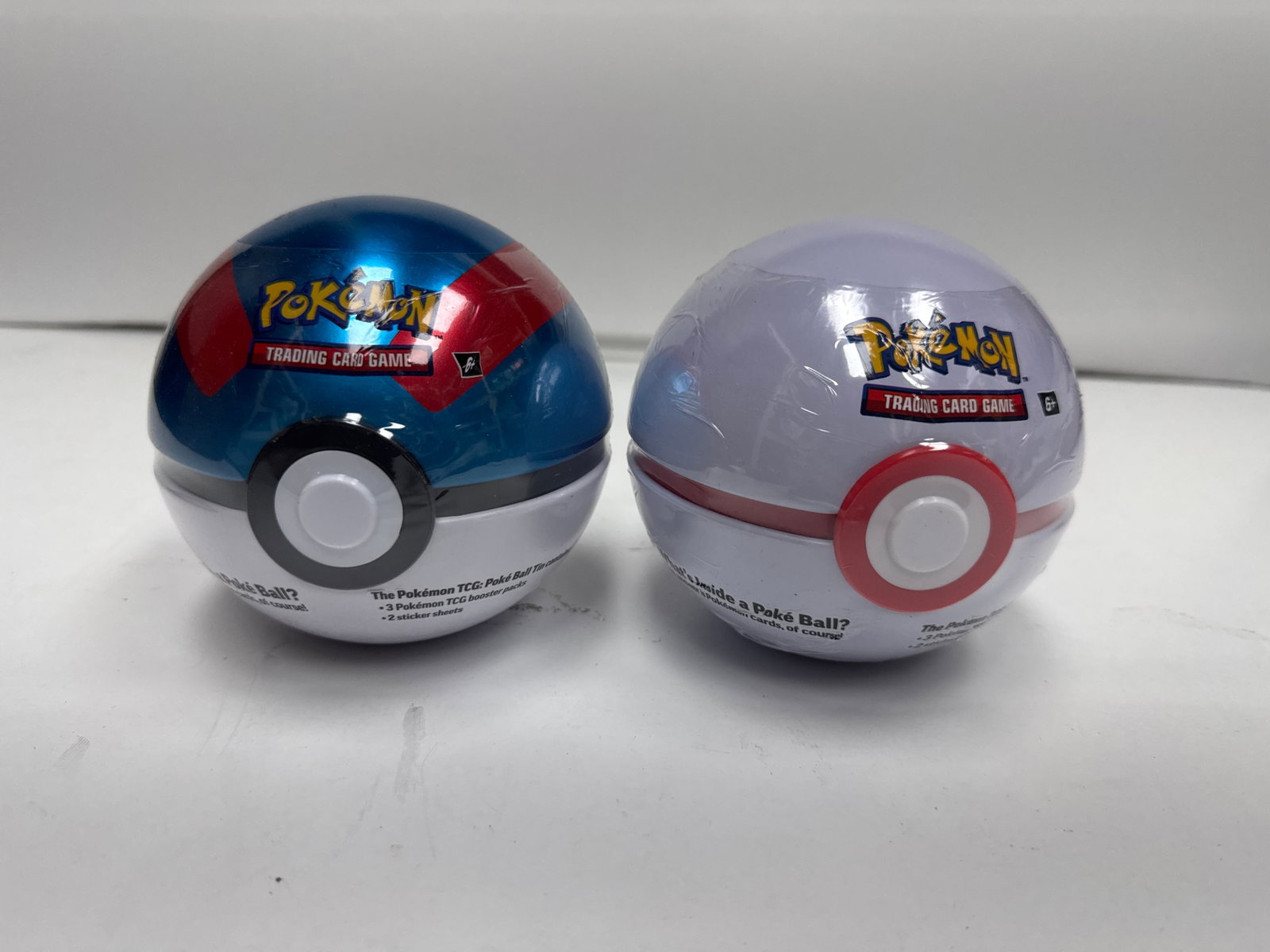 Pokemon Trading Card Game Poke Ball Tins (2-Pack): This lot includes two Pokemon Trading Card Game Poke Ball tins. Each tin contains 3 Pokemon TCG booster packs and 2 sticker sheets. One tin is blue and red, and the other is white and red.