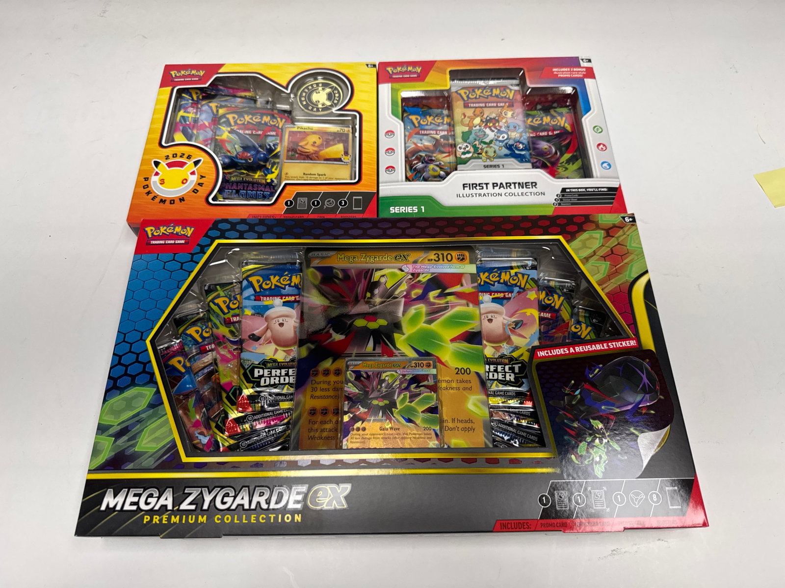 Pokemon TCG: Mega Zygarde EX Premium Collection, First Partner Illustration Collection & Pokemon Day: This lot includes three Pokemon Trading Card Game collections: the Mega Zygarde EX Premium Collection, featuring a Mega Zygarde EX card, a large lenticular card, and 8 booster packs, and the Firs