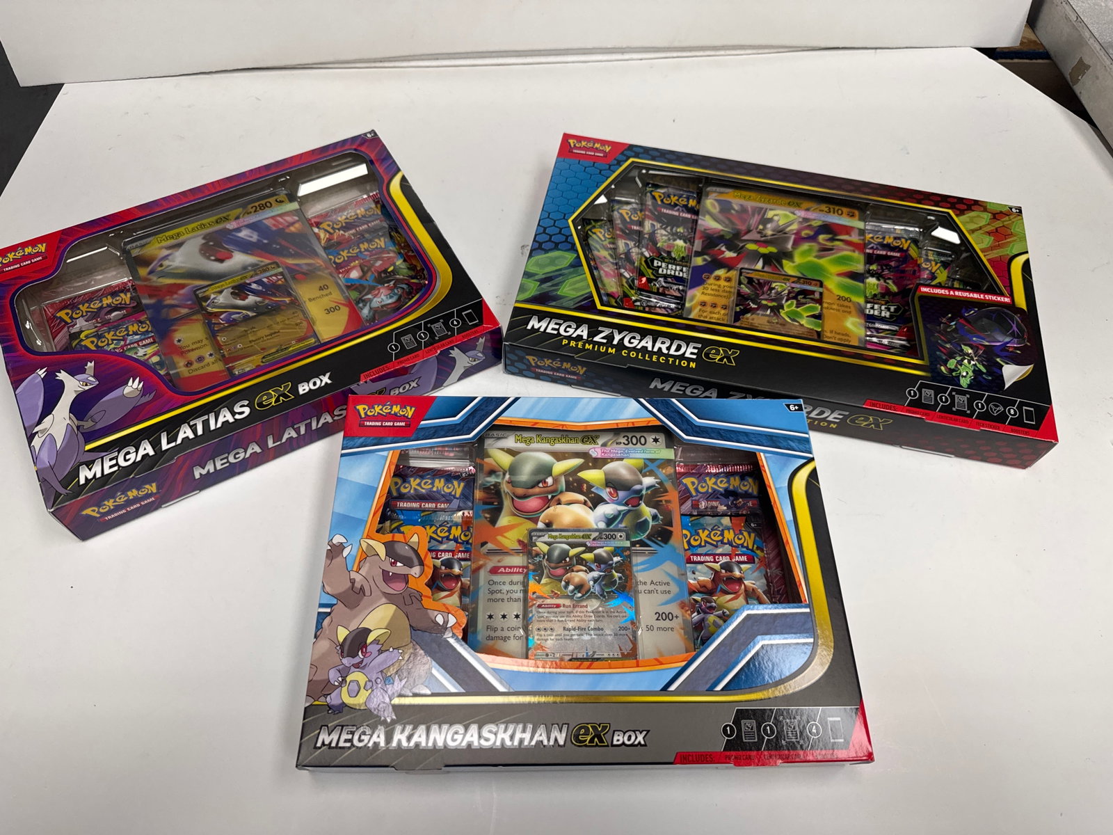 Pokemon TCG Mega Latias EX, Mega Zygarde EX, and Mega Kangaskhan EX Premium Collection Boxes: This lot includes three Pokemon Trading Card Game Premium Collection Boxes: Mega Latias EX, Mega Zygarde EX, and Mega Kangaskhan EX. Each box contains booster packs, special cards, and other collectib