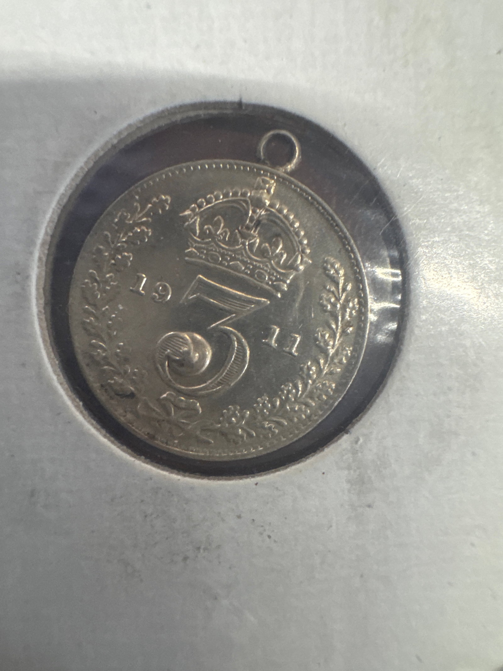1911 King George V Silver Three Pence Coin Pendant: A 1911 silver three pence coin featuring King George V on one side and a crown on the other. The coin has been modified into a pendant with a small turquoise stone accent and a loop for hanging.