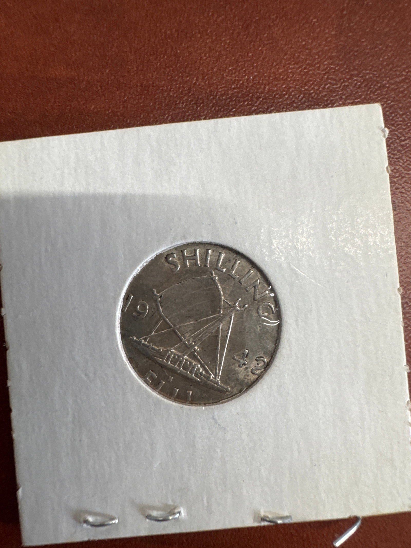 1942 East Africa Shilling Coin: A silver coin from East Africa, dated 1942. The obverse features a portrait of King George VI, and the reverse depicts a sailing ship. The coin is housed in a protective sleeve.