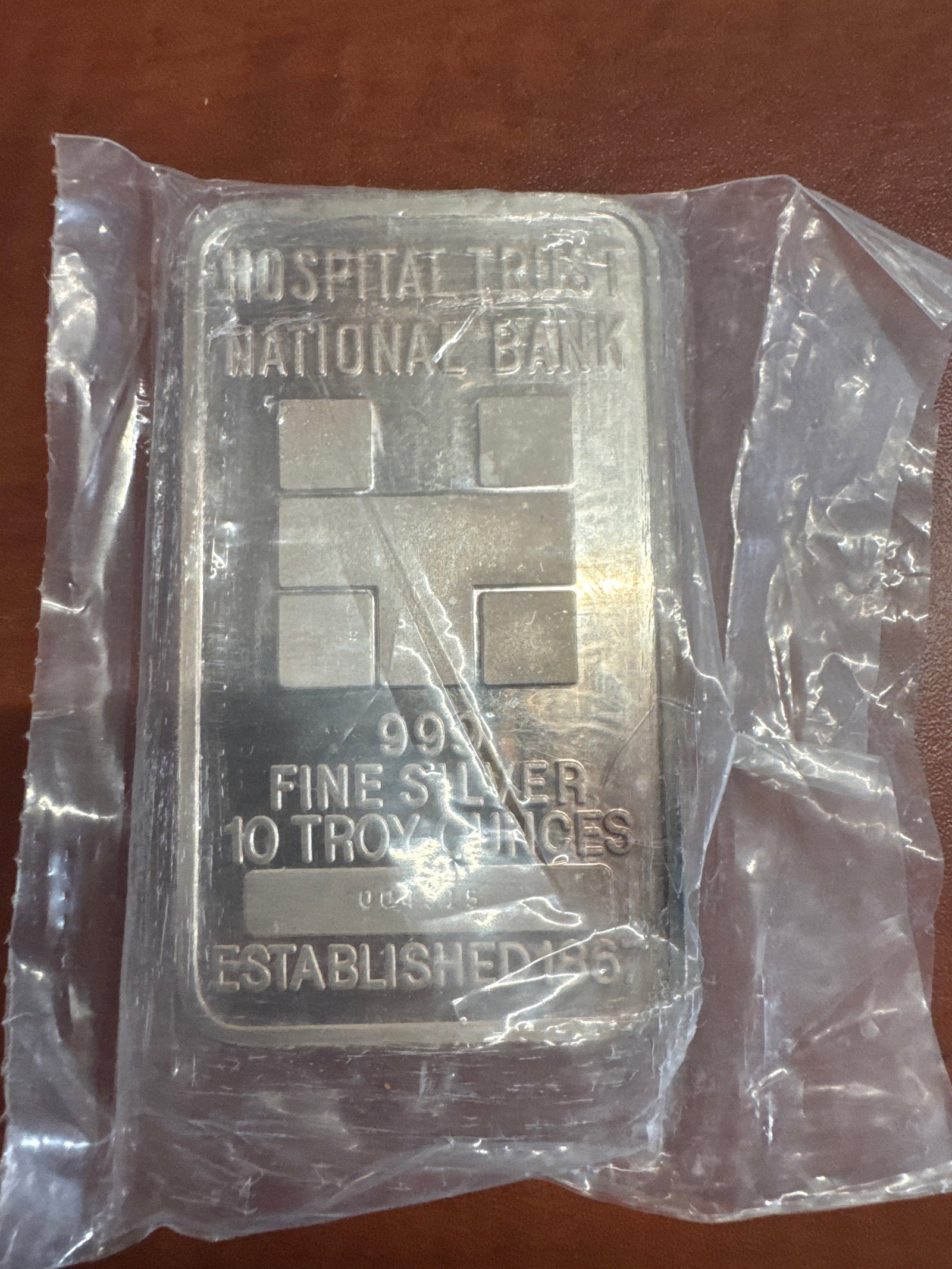 Hospital Trust National Bank 10 Troy Ounces 999 Fine Silver Bar: A 10 troy ounce bar of 999 fine silver from Hospital Trust National Bank, established in 1867. The bar features the bank's logo and details on the front, with a decorative pattern on the reverse. 