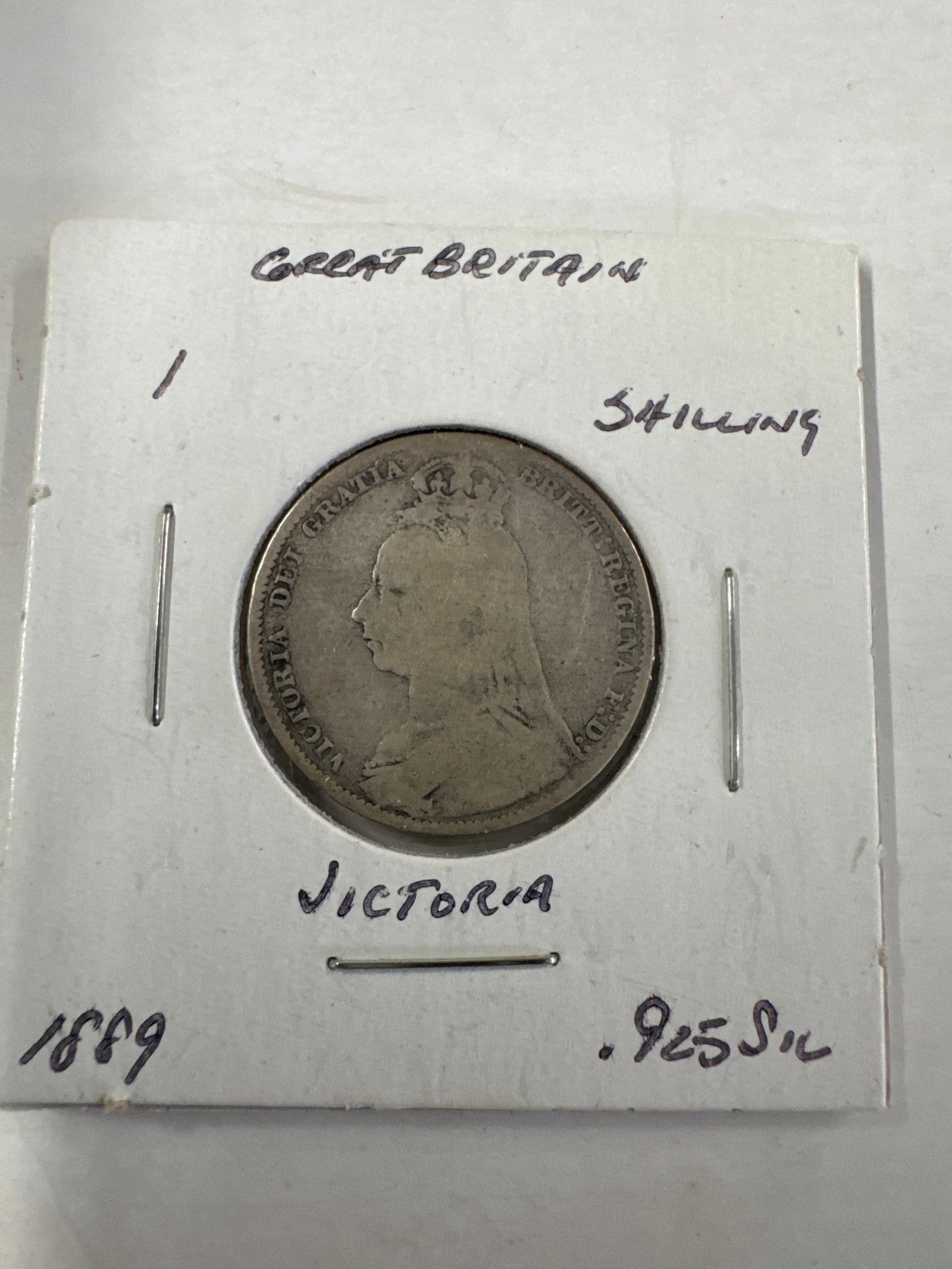 Great Britain 1889 Victoria Silver 1 Shilling Coin: A silver 1 shilling coin from Great Britain, dated 1889, featuring Queen Victoria's profile on the obverse and the royal coat of arms on the reverse. The coin is composed of .925 silver.