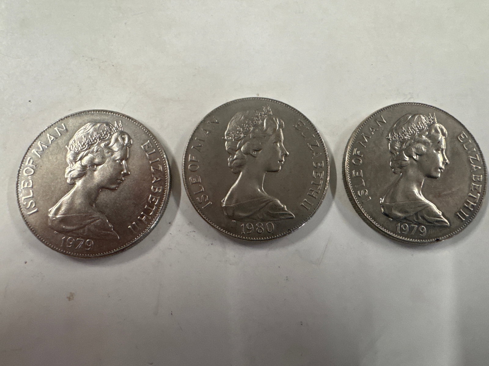 Three Isle of Man One Crown Coins: A collection of three Isle of Man One Crown coins. Two coins are dated 1979 and feature a profile of Queen Elizabeth II. The third coin is dated 1980 and also features Queen Elizabeth II. 