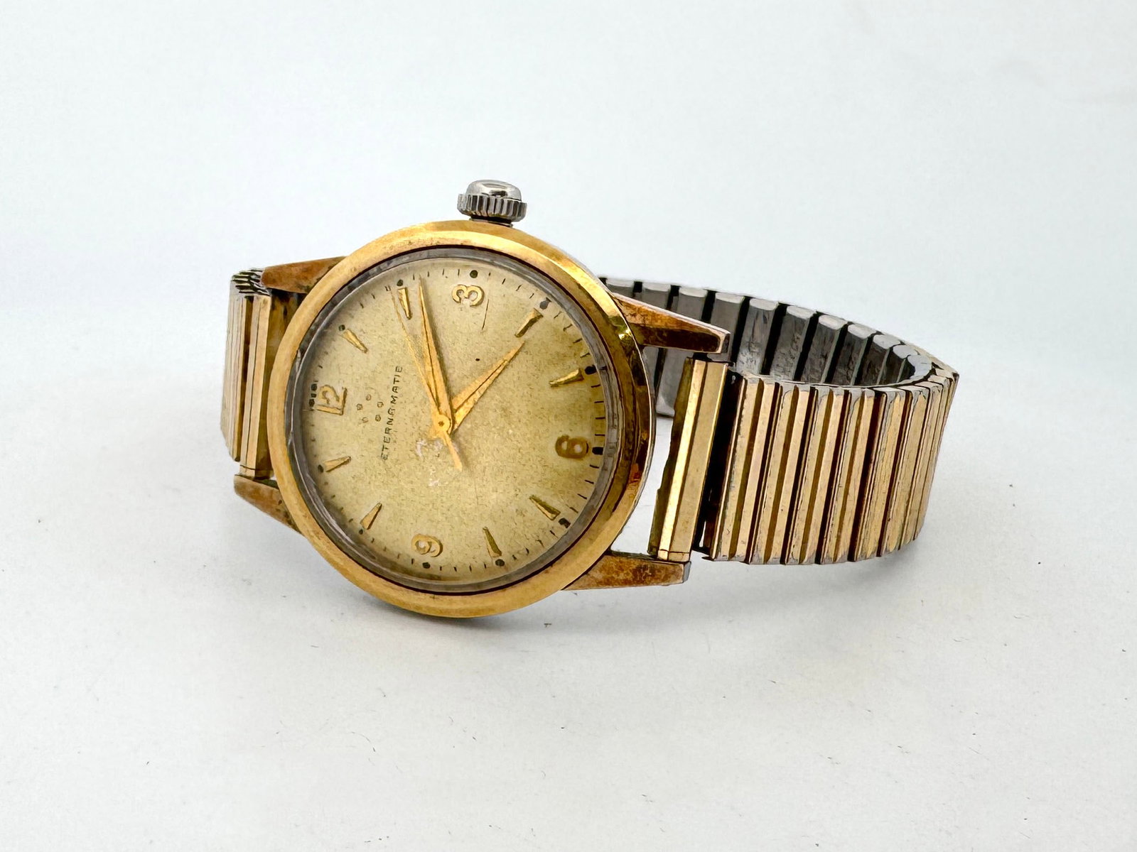 Eterna Matic Vintage Men's Watch: A vintage Eterna Matic men's wristwatch featuring a gold-tone case and dial with Arabic numerals and markers. The watch has a stainless steel case back and an expandable metal bracelet. The moveme