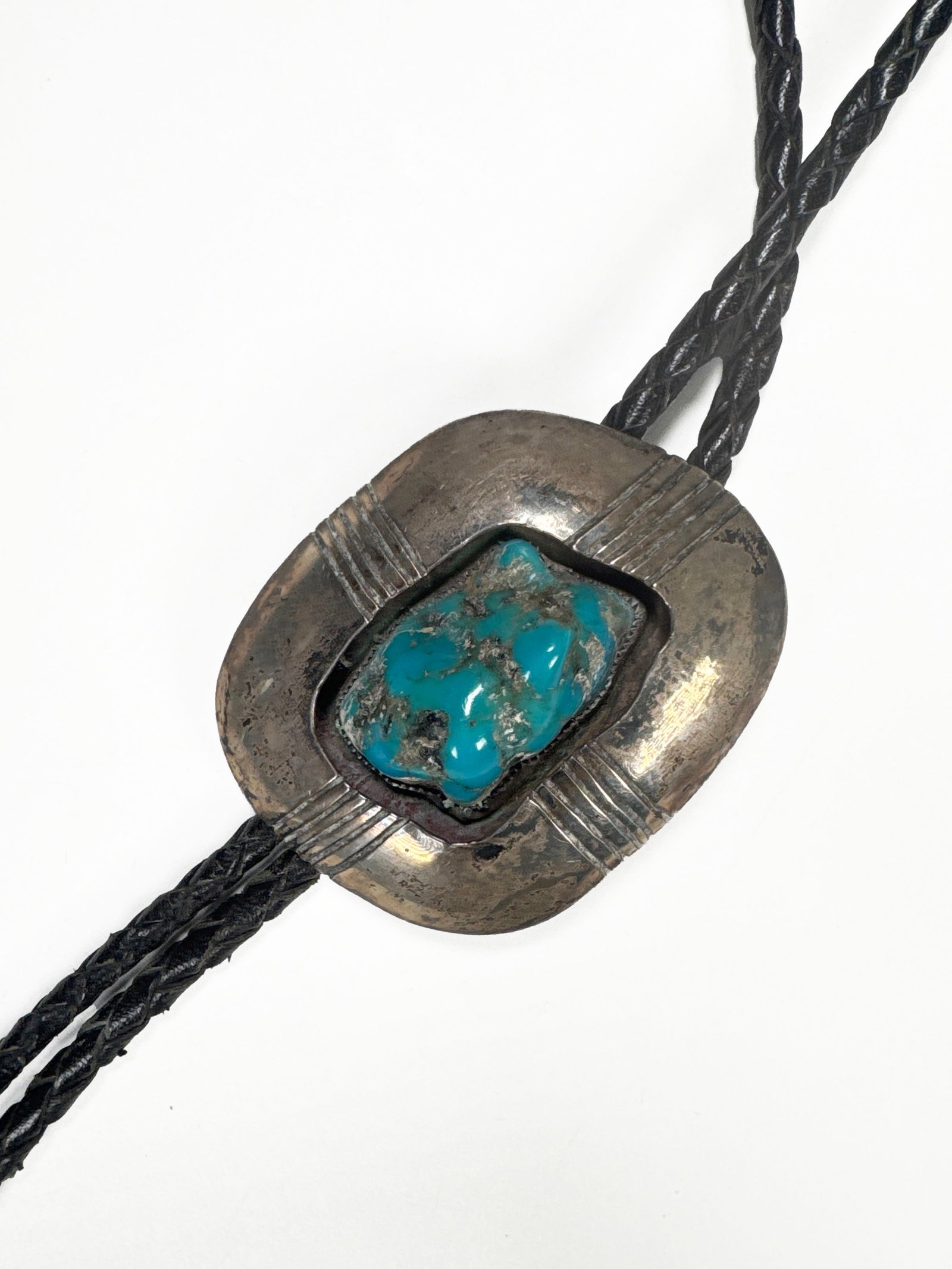 Ray Bennet Sterling Silver & Turquoise Bolo Tie: A vintage sterling silver bolo tie featuring a central turquoise stone. The silver setting has a textured, rectangular design with grooved lines. The braided leather cord has metal tips. The back is m