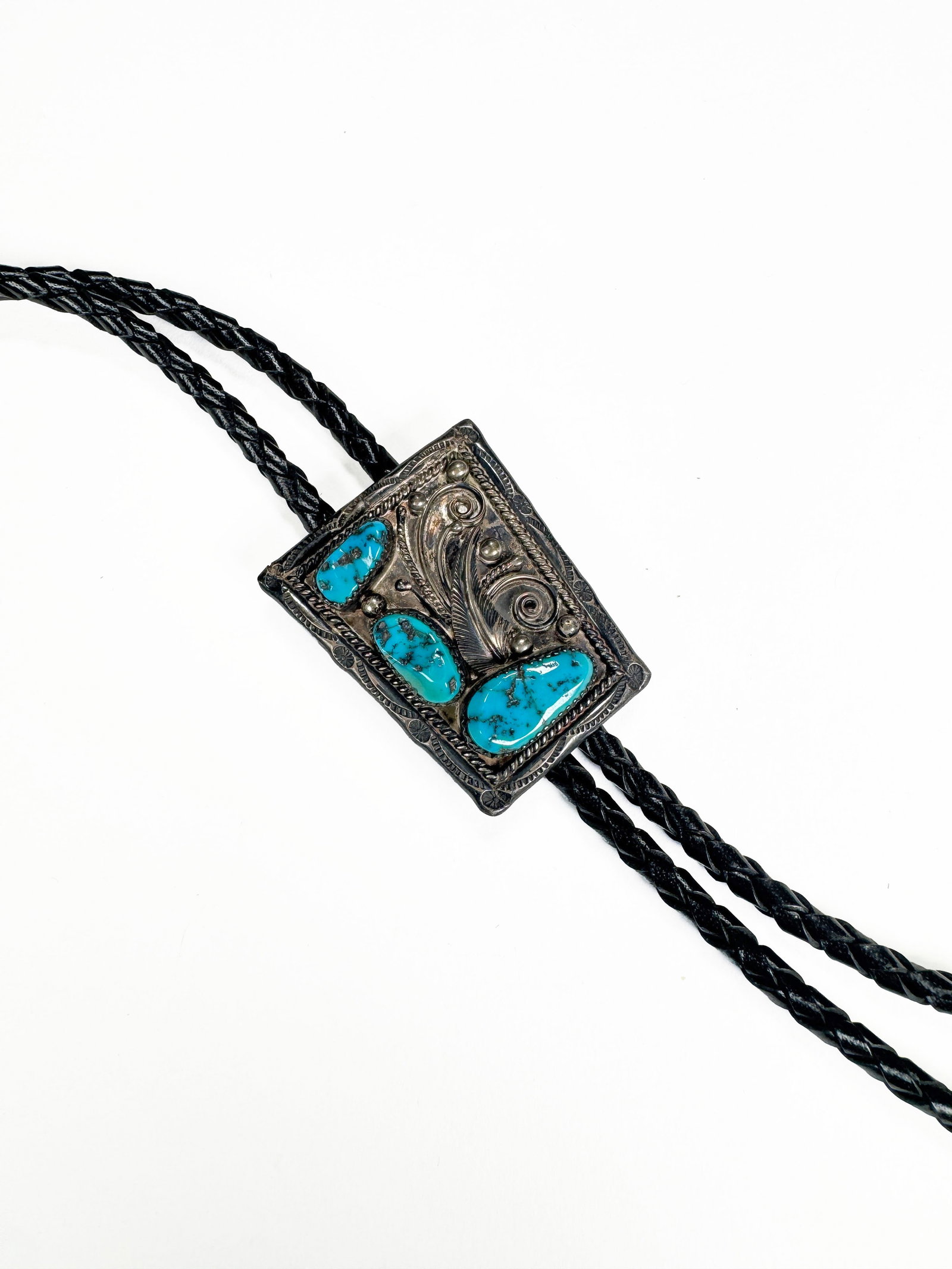 Sterling Silver & Turquoise Signed Bolo Tie: A vintage silver tone bolo tie featuring a rectangular centerpiece adorned with polished turquoise stones. The silver has intricate scrollwork and detailing. The tie has a braided black leather cord w