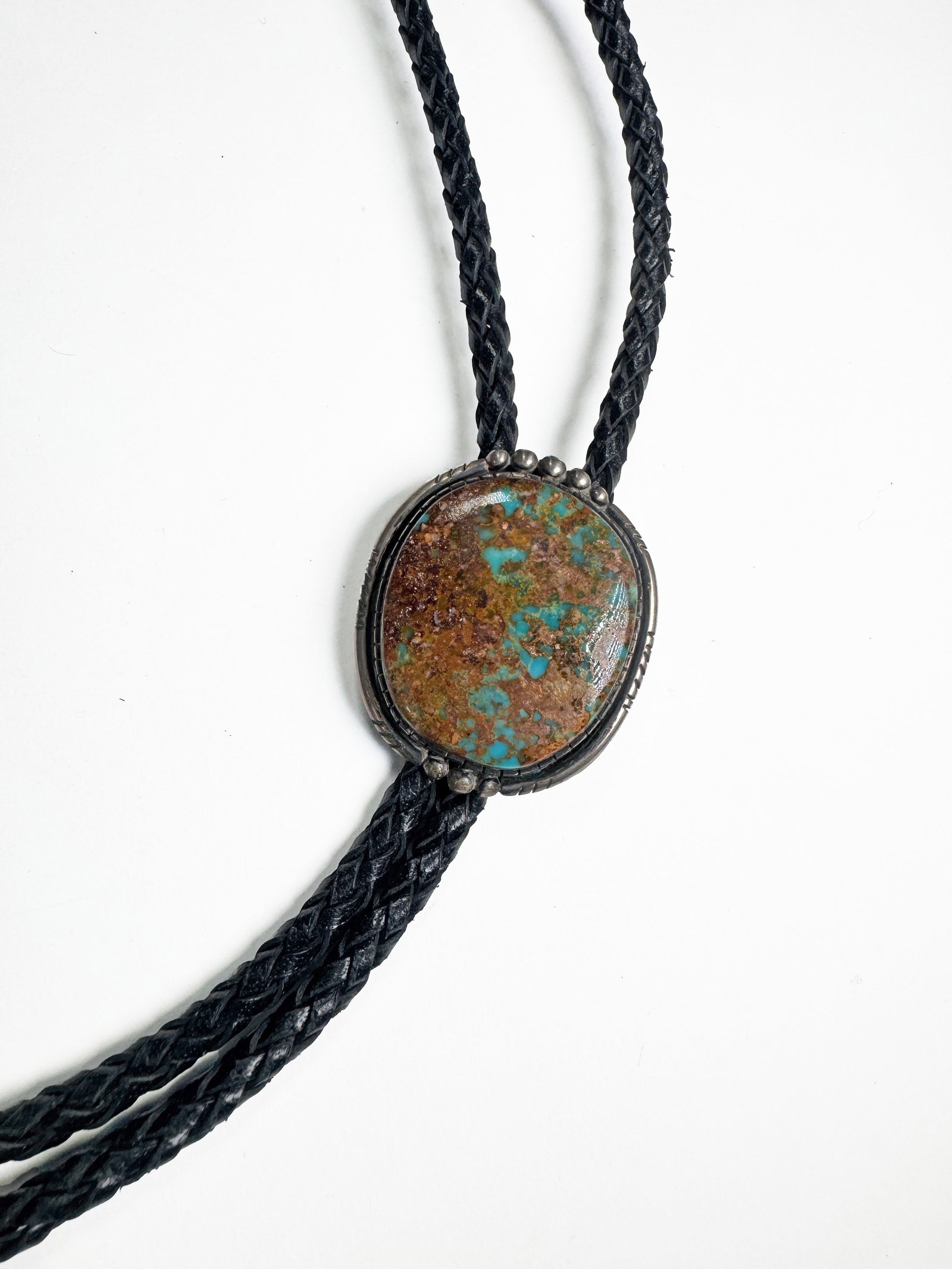 Sterling Silver Turquoise Signed Bolo Tie: A vintage bolo tie featuring a central oval-shaped turquoise stone with brown and blue matrix, set in a decorative silver-tone bezel. The tie has a braided black leather cord with silver-tone tips. Si