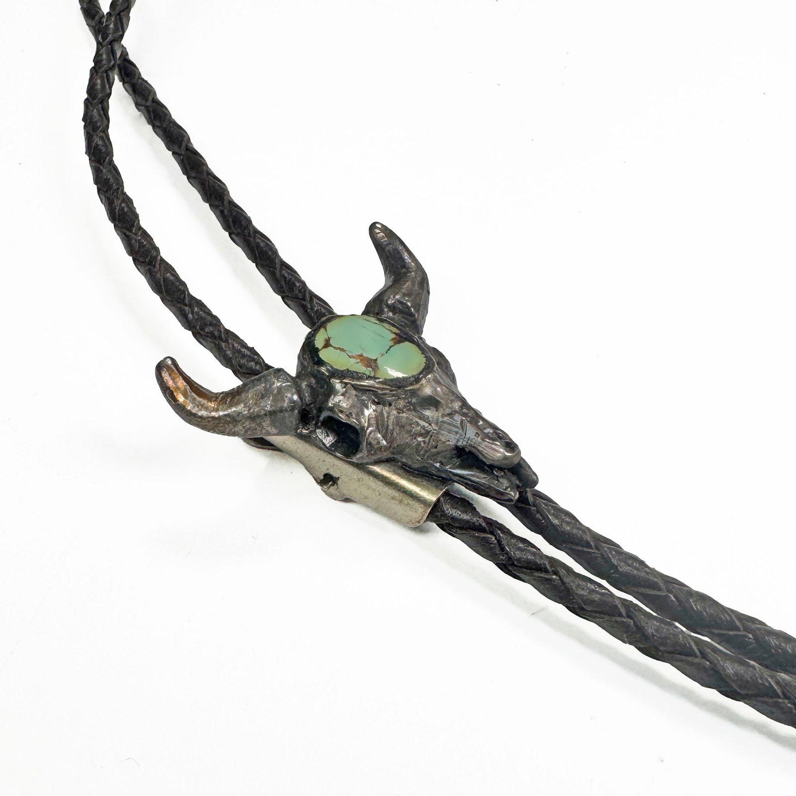 Sterling Silver & Turquoise Bull Bolo Tie: A striking bolo tie featuring a detailed silver tone bull skull pendant with a central turquoise stone. The pendant is suspended on a braided black leather cord with silver-tone bolo tips. Size: 18.5&