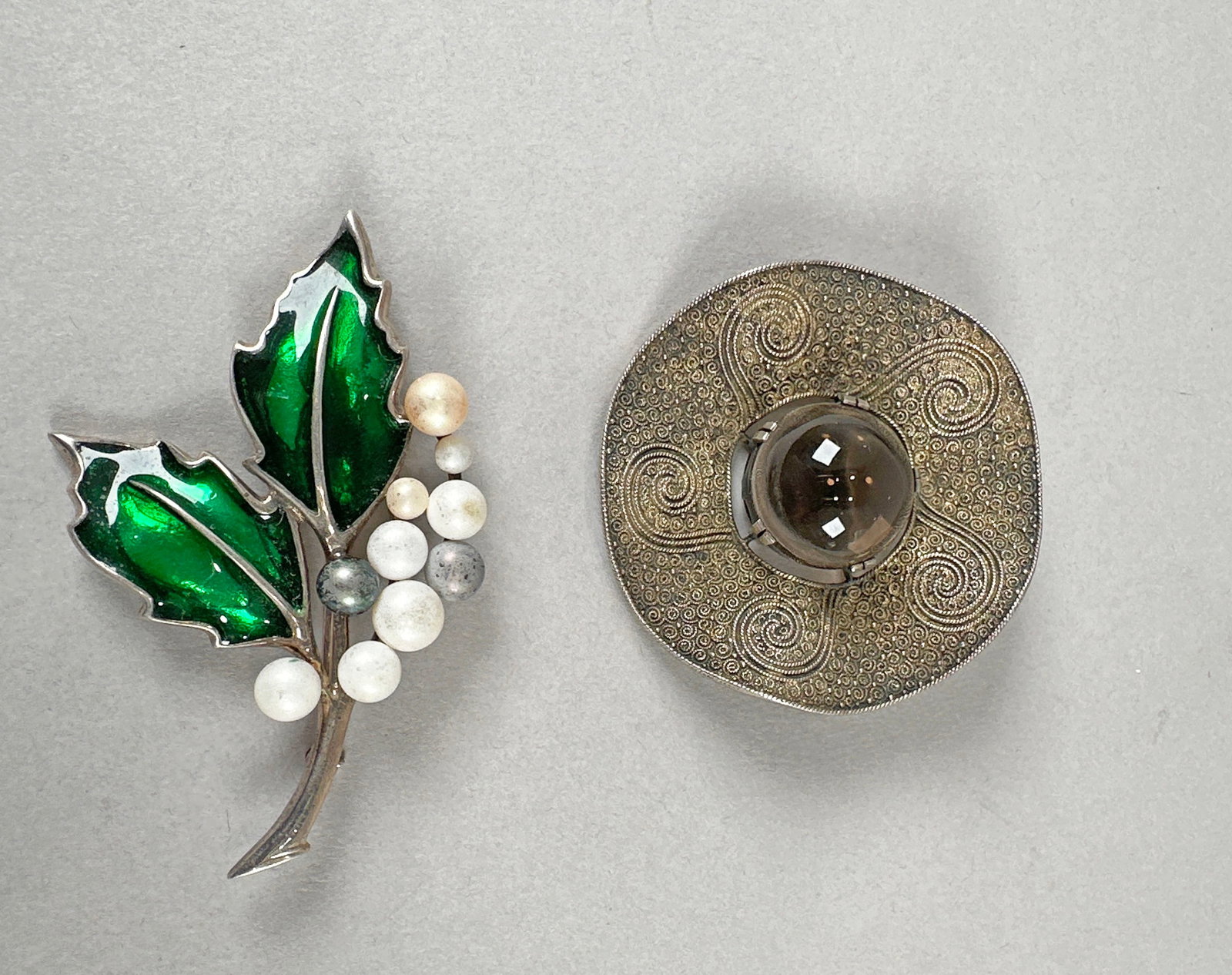 Two Sterling Silver Brooches: This lot features two sterling silver brooches. The first is a circular brooch with intricate filigree work and a central smoky quartz stone. The second is a charming holly berry design with green ena