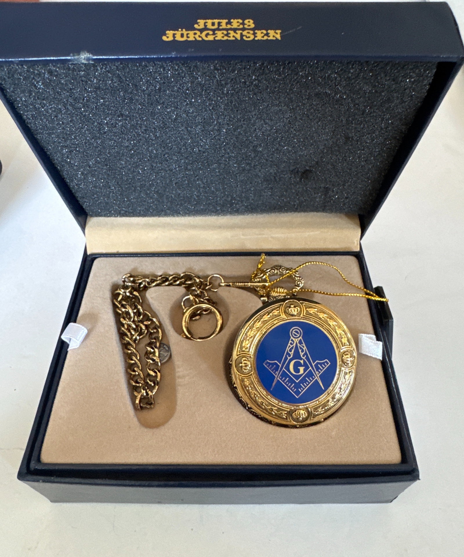 Jules Jurgensen Gold-Tone Masonic Pocket Watch with Chain: A gold-tone pocket watch by Jules Jurgensen, featuring a Masonic emblem on the cover and a quartz movement. The watch comes with its original presentation box and a matching chain. The case is made