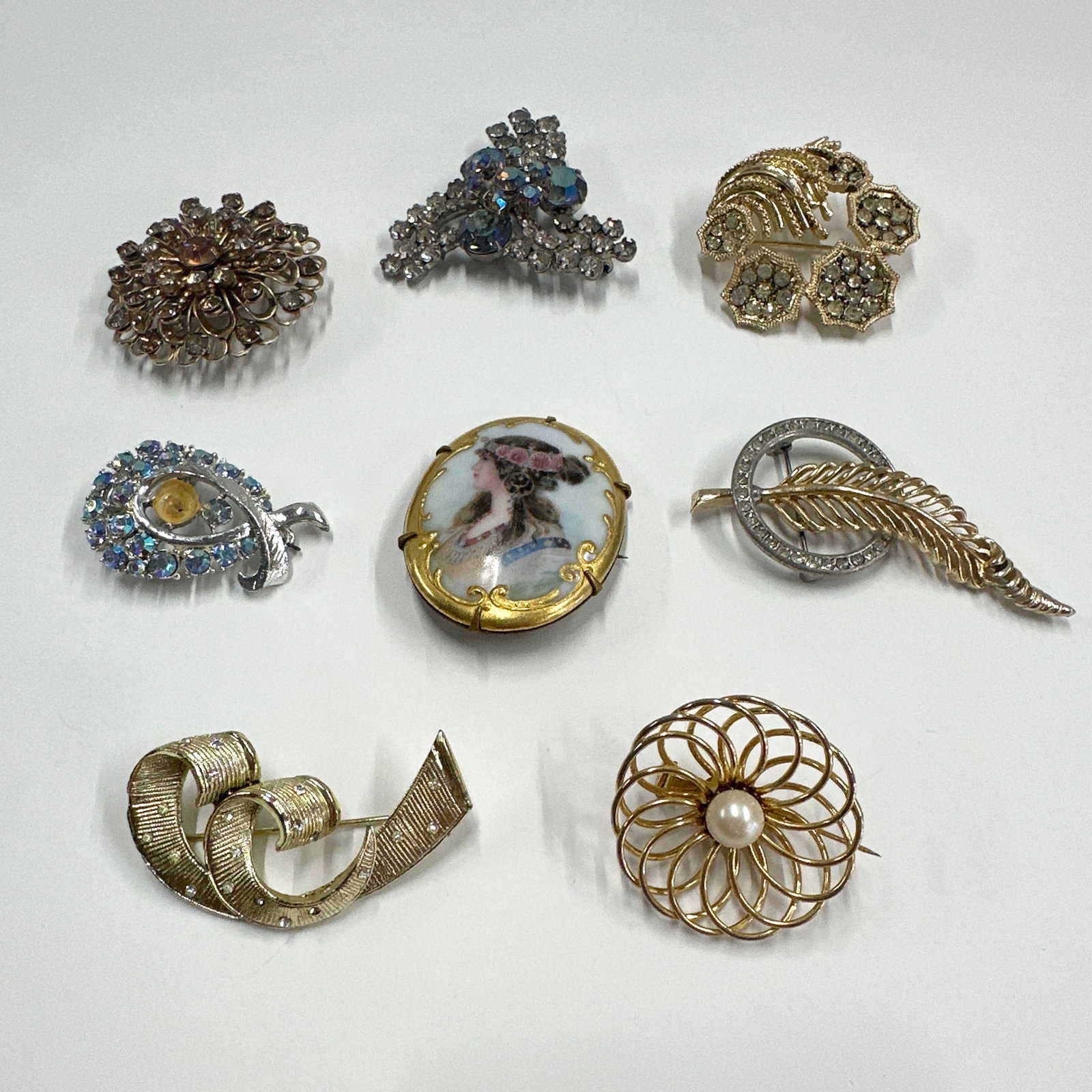 Collection of Vintage Brooches: A collection of eight vintage brooches, featuring various designs including floral motifs, a feather, a cameo, and abstract shapes. Some brooches are adorned with rhinestones and pearls. The total wei